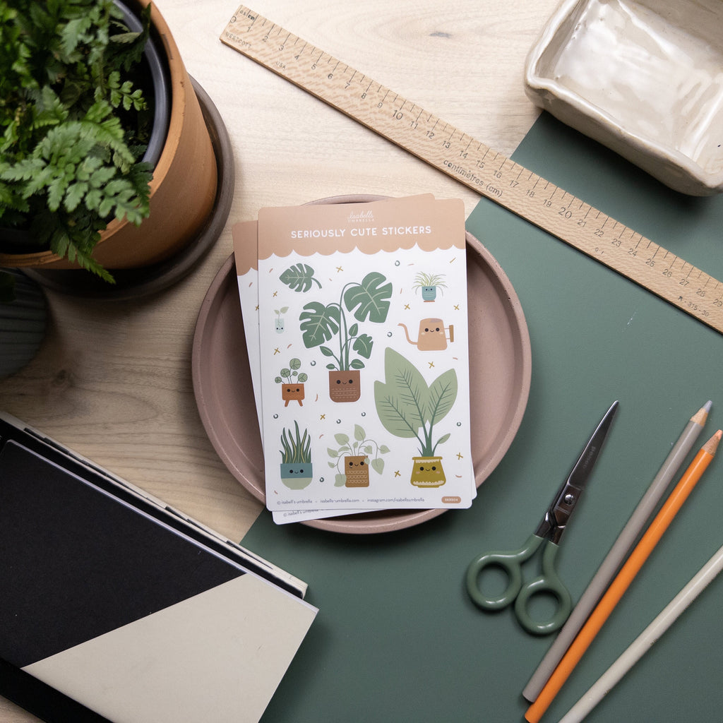 Vinyl sticker set featuring nine different illustrated potted plants photographed on top of a pink tray surrounded by miscellaneous art supplies.
