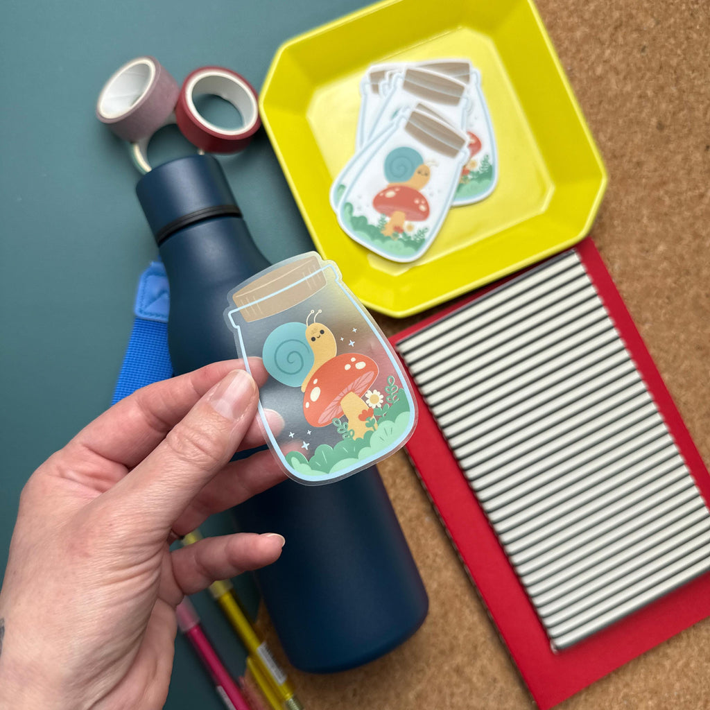 A hand holding a cute sticker featuring a yellow snail sitting on a red mushroom inside a jar. The background includes a blue water bottle, red and pink washi tape rolls, yellow tray with stickers, and a notebook partially visible.