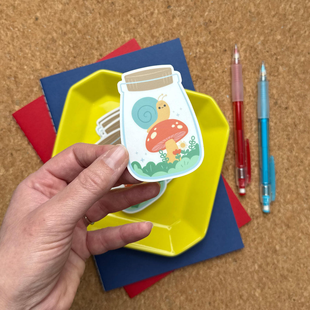 A close-up of a hand holding a sticker showing a snail on a red mushroom inside a jar, surrounded by plants and flowers.