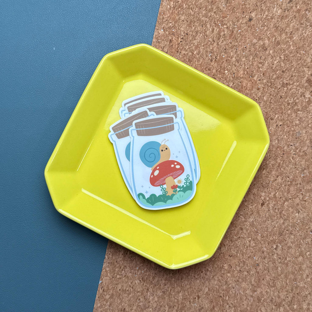 A stack of stickers featuring the yellow snail on a red mushroom inside a jar, neatly placed in a bright yellow tray. The background includes a corkboard and a dark blue surface.