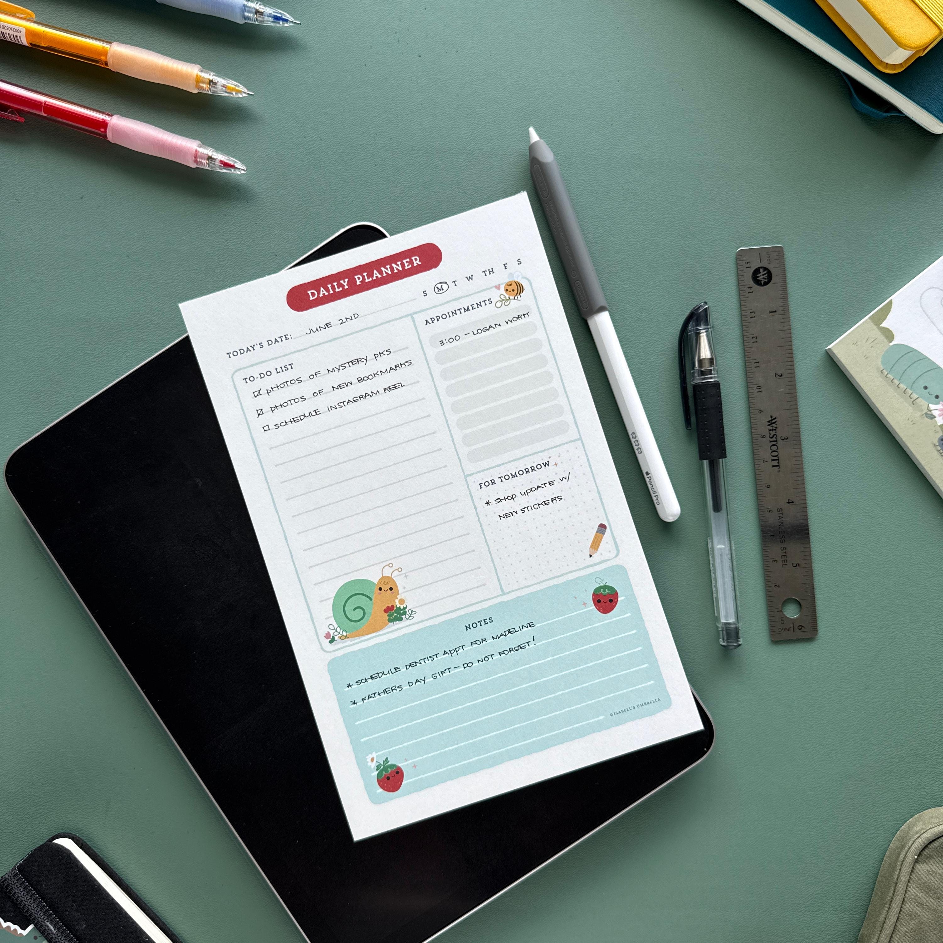 A flat lay of a cute daily planner notepad featuring whimsical illustrations of a snail and ladybugs, placed on top of a black tablet on a green desk. Surrounding the notepad are colorful pens, a white stylus, a black gel pen, and a metal ruler.