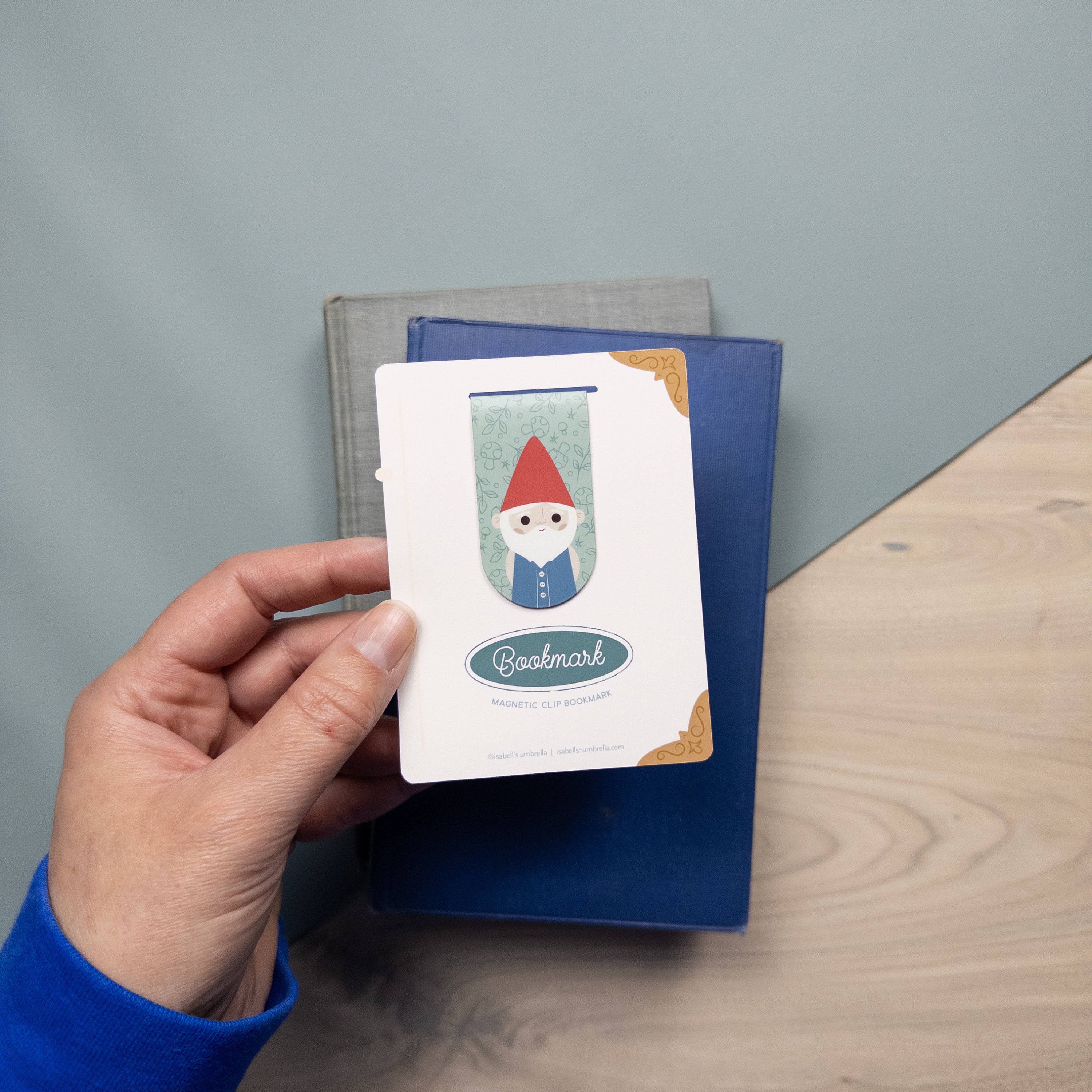 Hand holding a packaged magnetic bookmark of a gnome with stacked books in the background