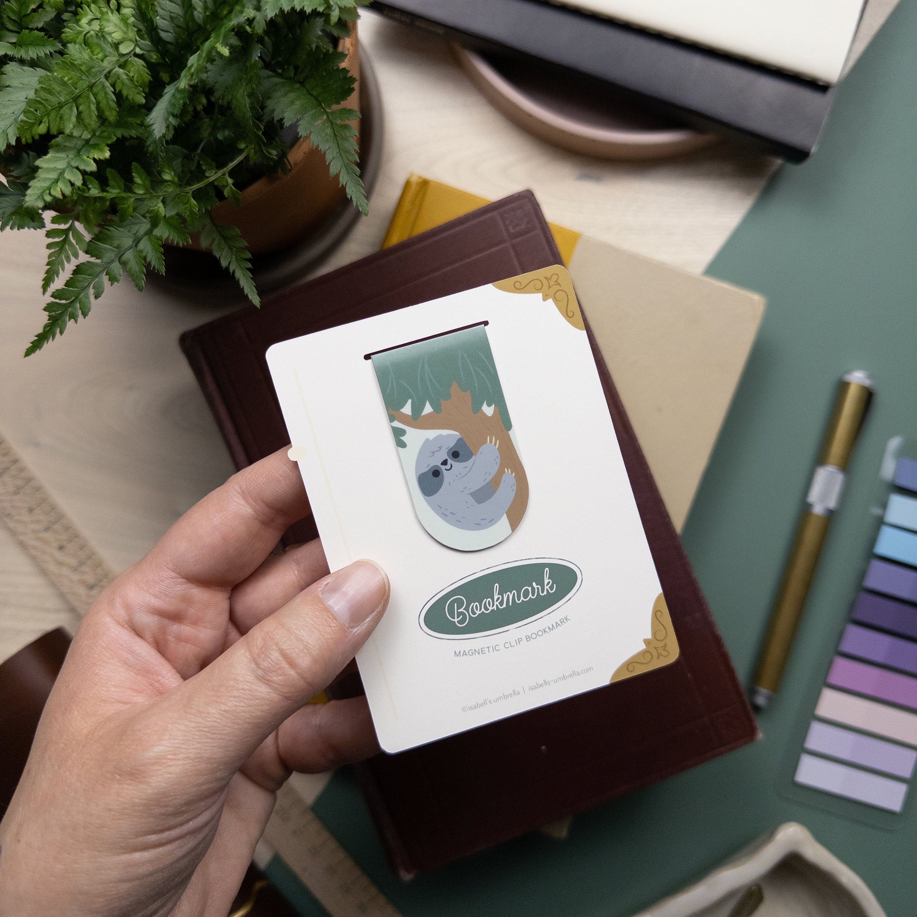 Hand holding a packaged magnetic clip bookmark featuring an illustrated sloth in a tree.