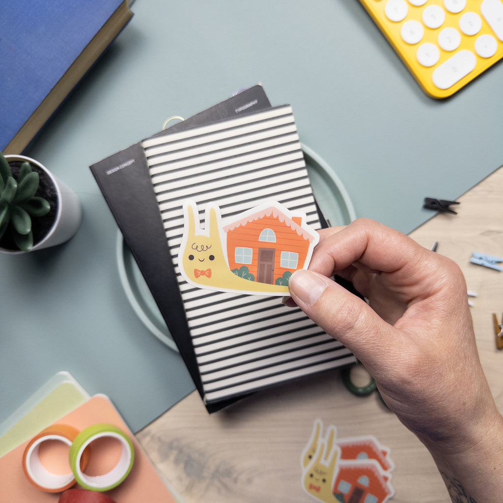 Hand holding an illustrated vinyl sticker of a snail with a house for a shell.