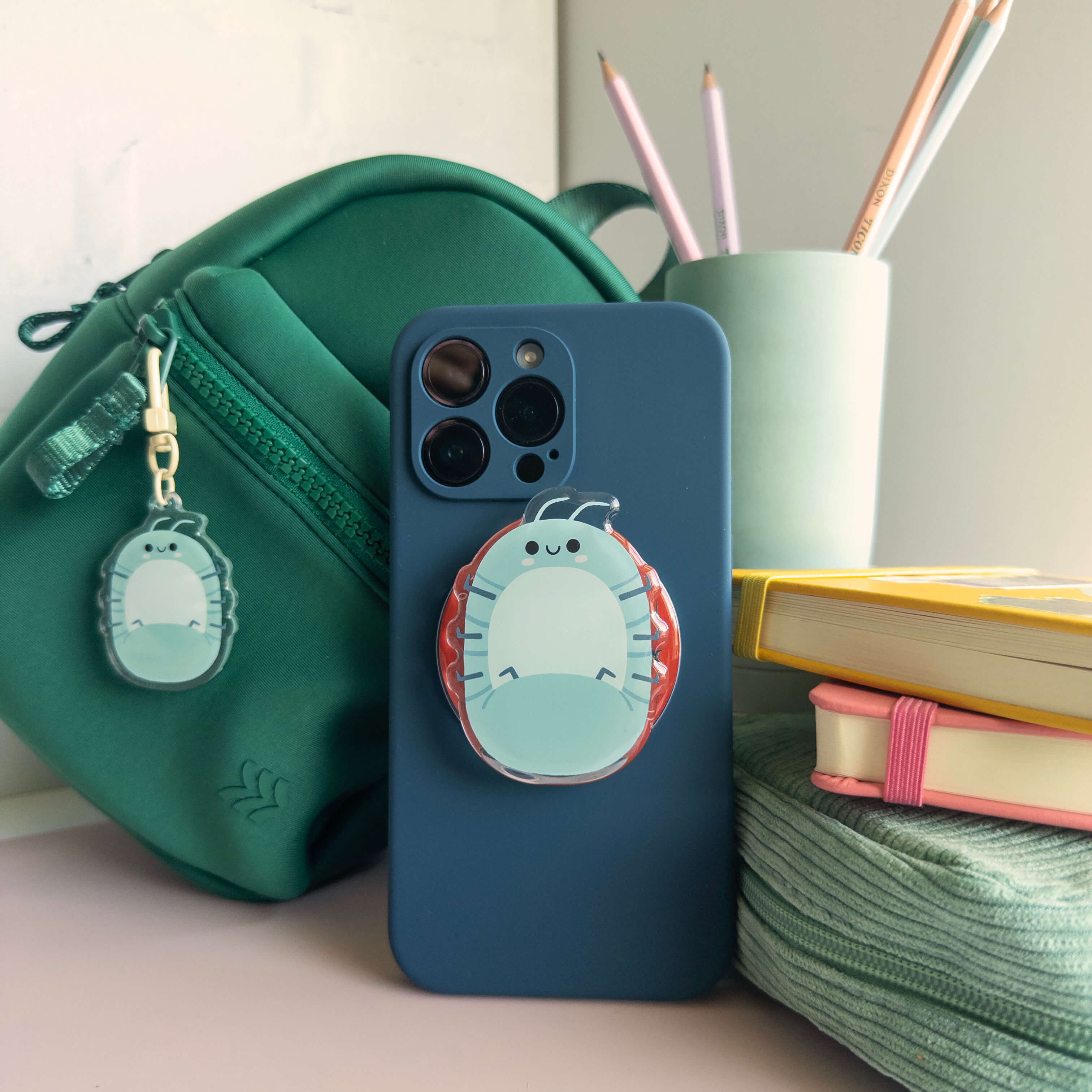 Phone case with a cute roly poly designed magnetic phone grip on a desk with a green bag and stationery items.