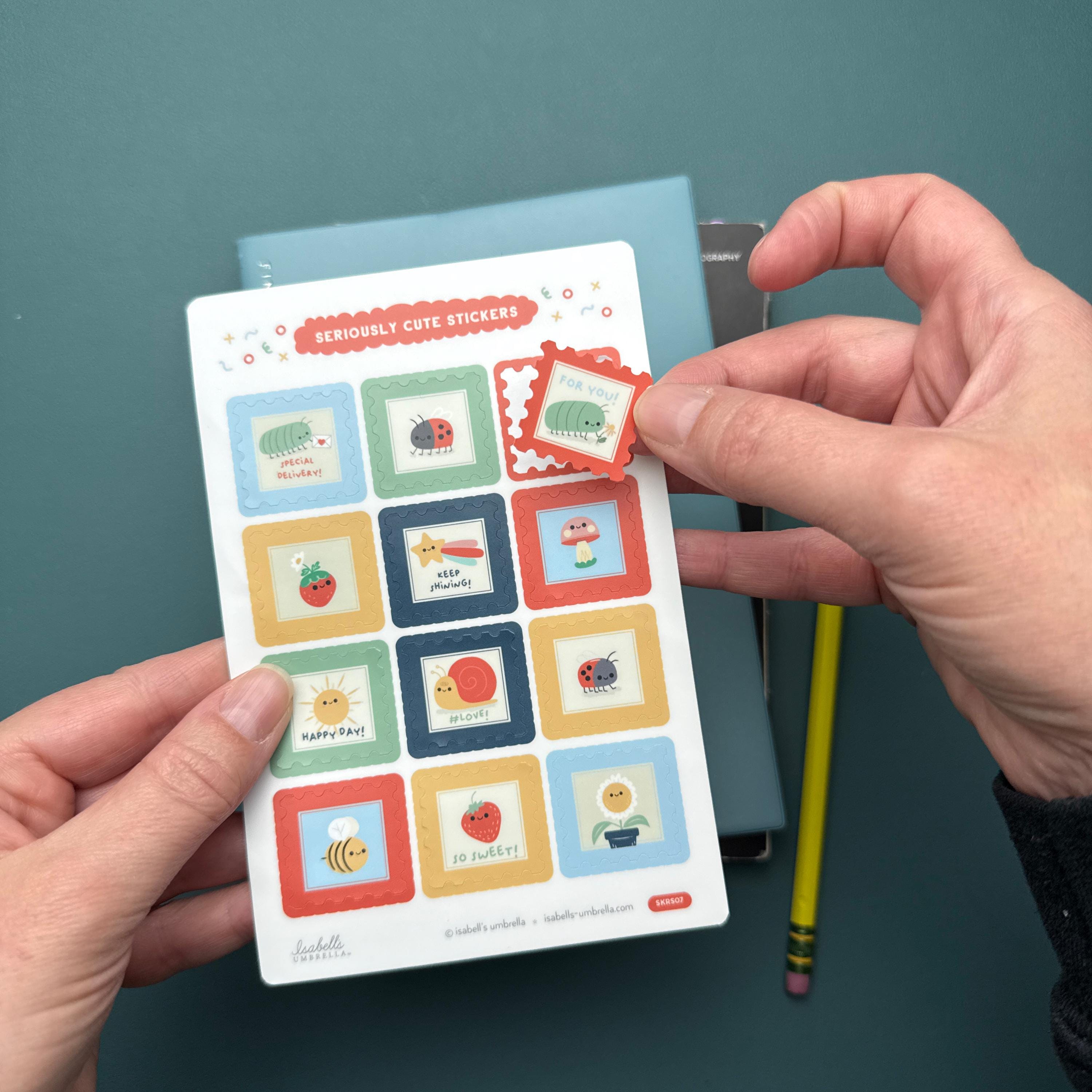 Hand peeling off a sticker with a roly-poly holding a flower, from a sheet of other stickers featuring bugs, flowers, and a sunshine.