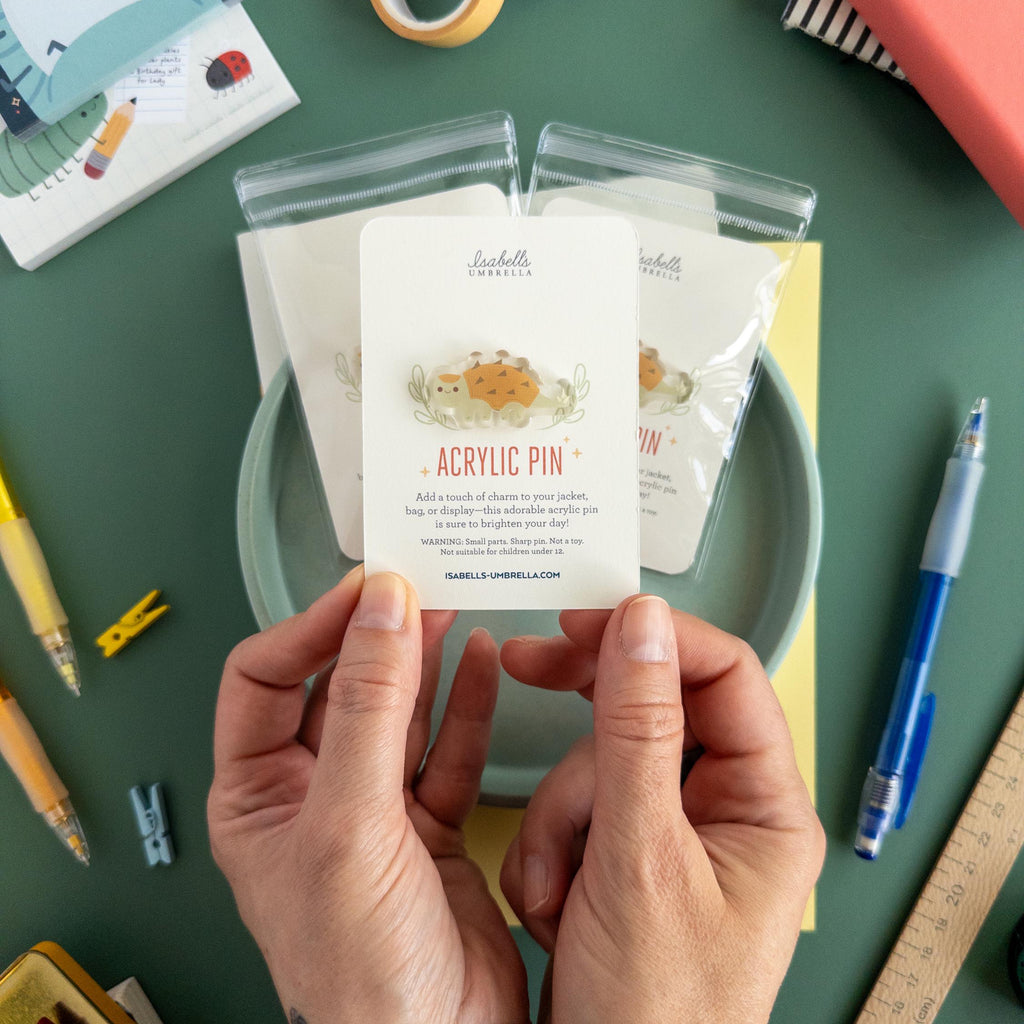 Person holding a cute Ankylosaurus acrylic pin with glossy epoxy finish with stationery items on a green surface