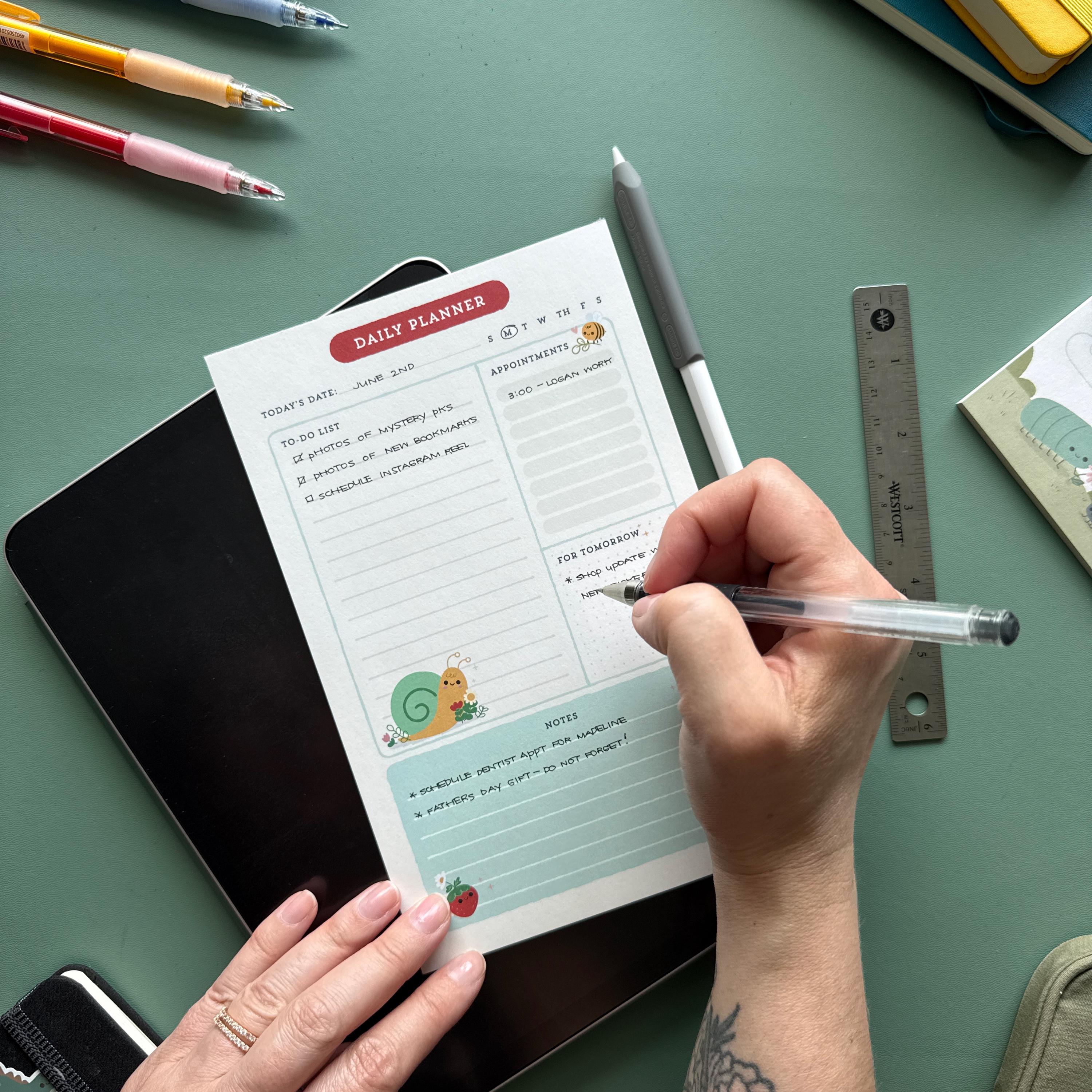 A person writes on a daily planner notepad with a black gel pen. The notepad is decorated with playful illustrations and has sections for to-do lists, appointments, and notes. Various stationery items, including pens and a ruler, are arranged on the desk.