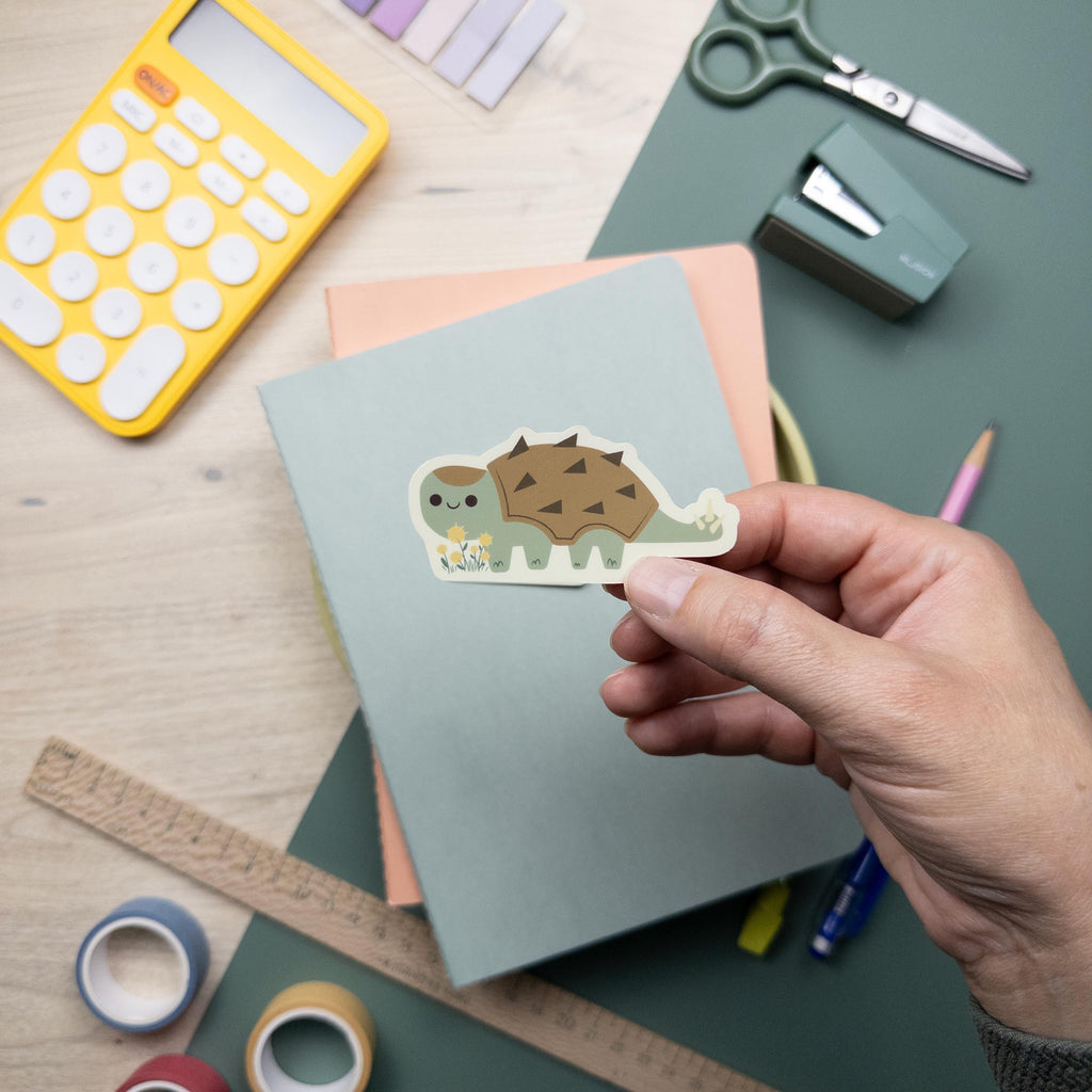 Hand holding a cute Ankylosaurus vinyl sticker with matte finish, kawaii dinosaur waterproof decal for laptops, water bottles, and journals.