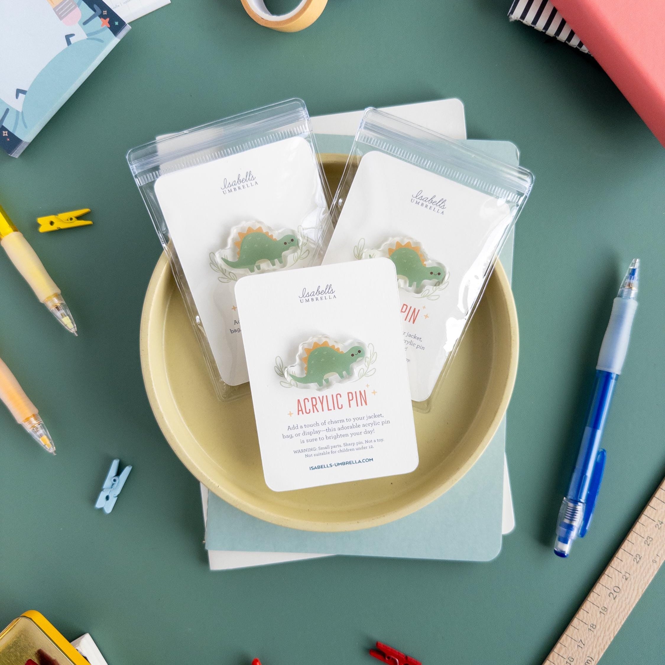 Three packaged adorable Stegosaurus pins arranged on a yellow plate surrounded by a variety of other art supplies.