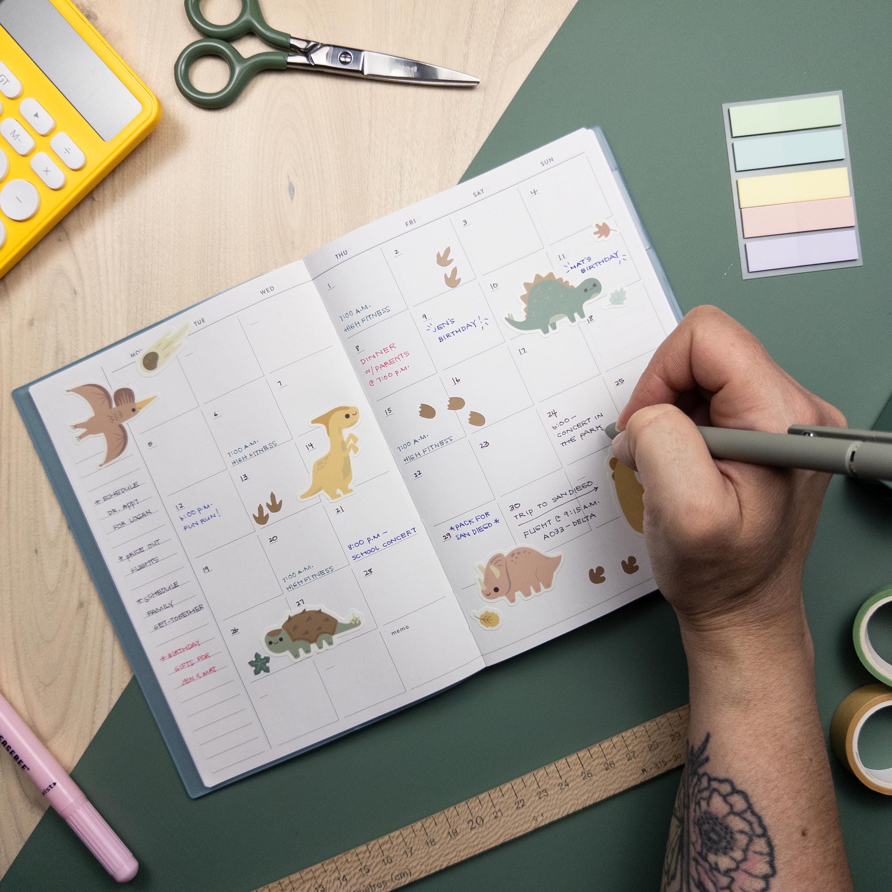 hand holding a pen and writing on a calendar decorated with a variety of dinosaur themed stickers.