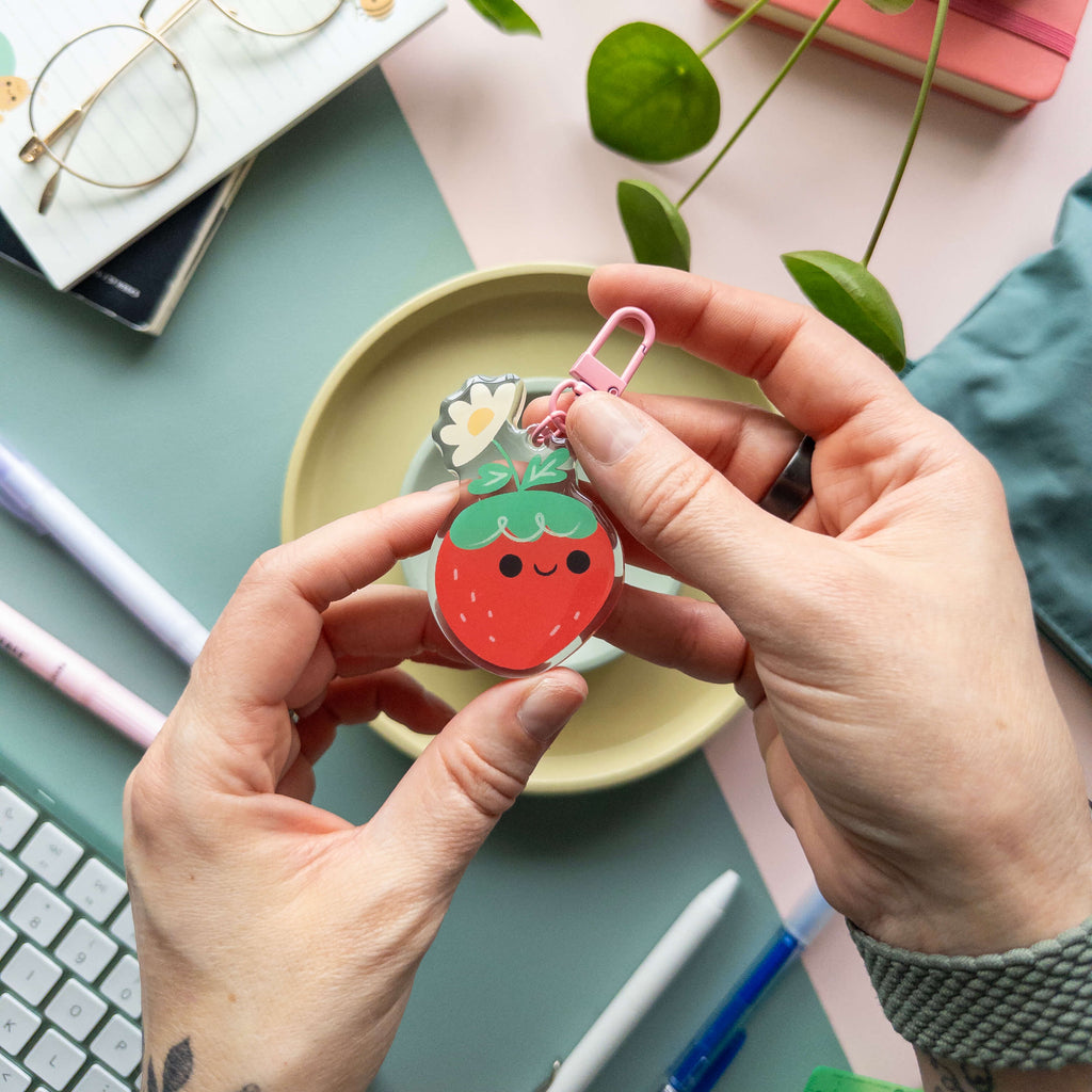 Person holding a strawberry-shaped acrylic keychain over a desk with stationery items.
