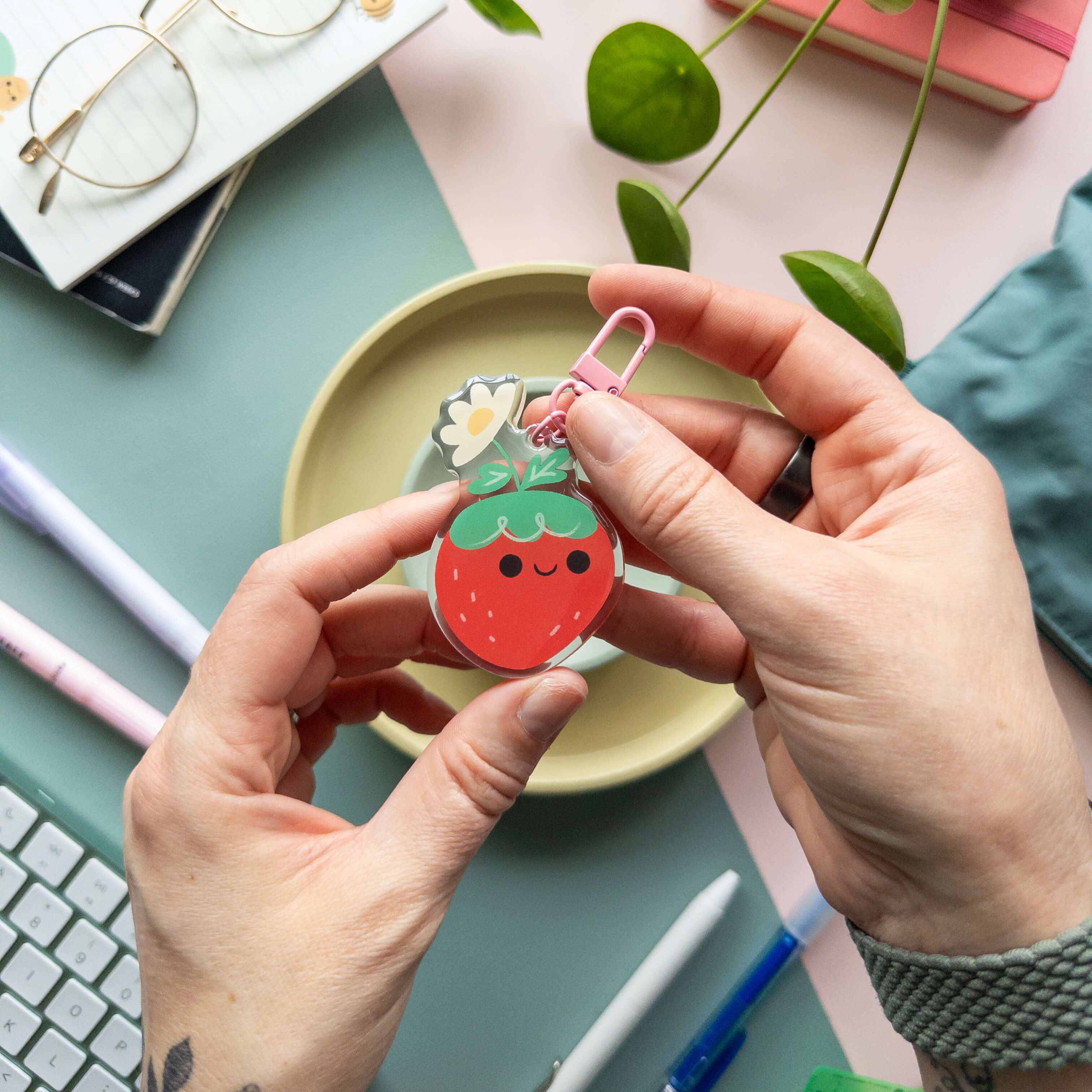 Person holding a strawberry-shaped acrylic keychain over a desk with stationery items.