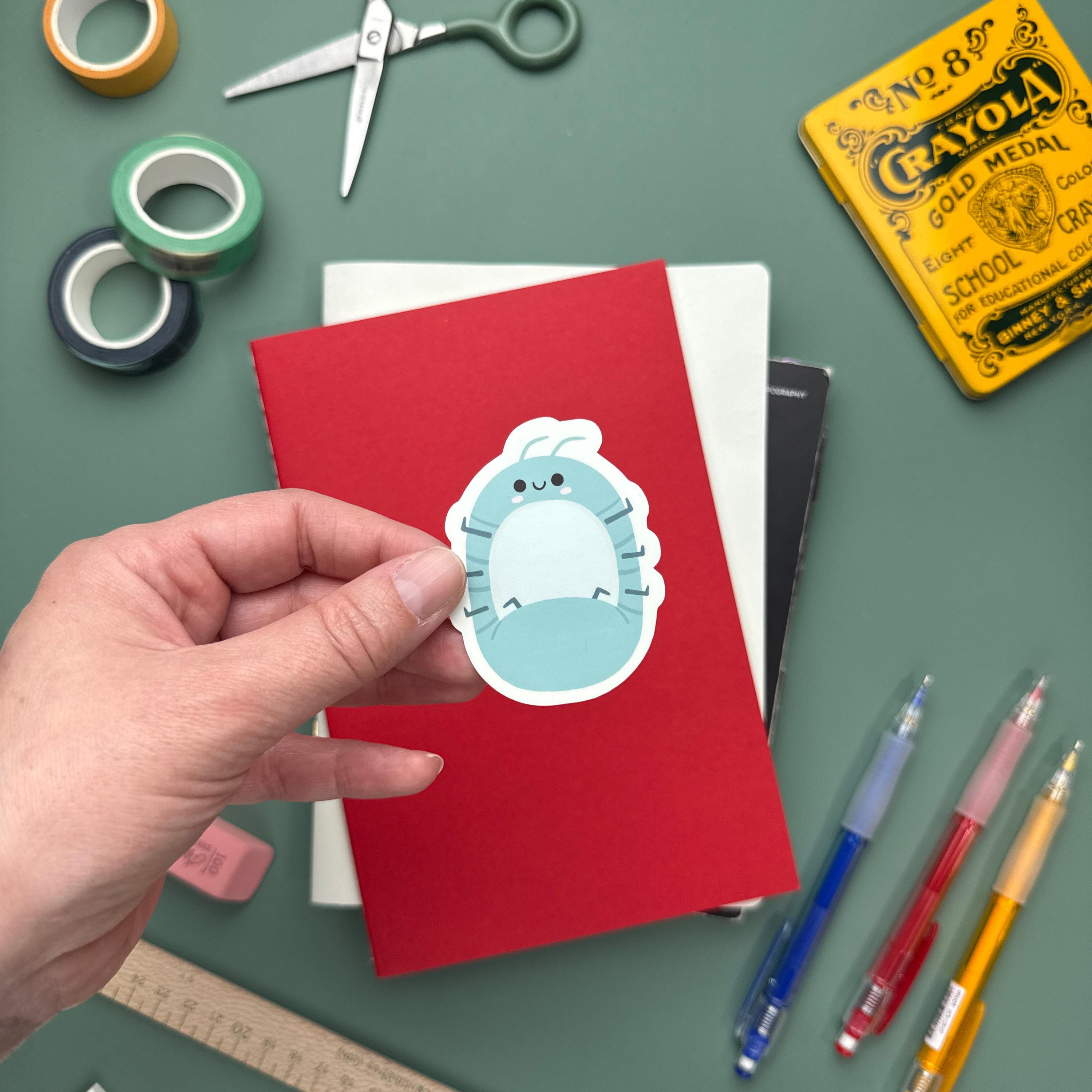 Hand holding a kawaii bug sticker featuring a pill bug. Surrounded by colorful stationery including washi tape, scissors, and notebooks—ideal for journals, planners, and scrapbooking.