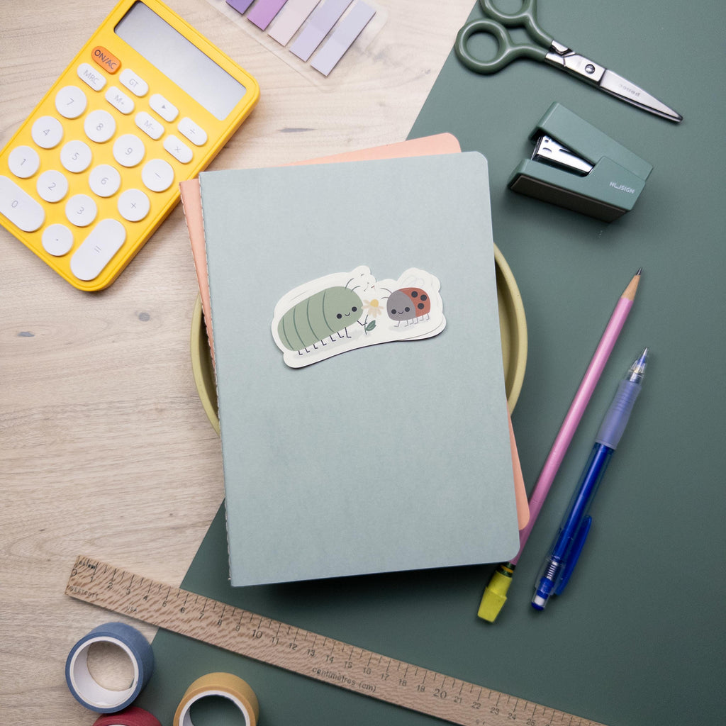 Photo of an illustrated sticker of a roly poly holding a flower and a lady bug. Sticker is set a top a stack of notebooks surrounded by miscellaneous art supplies.