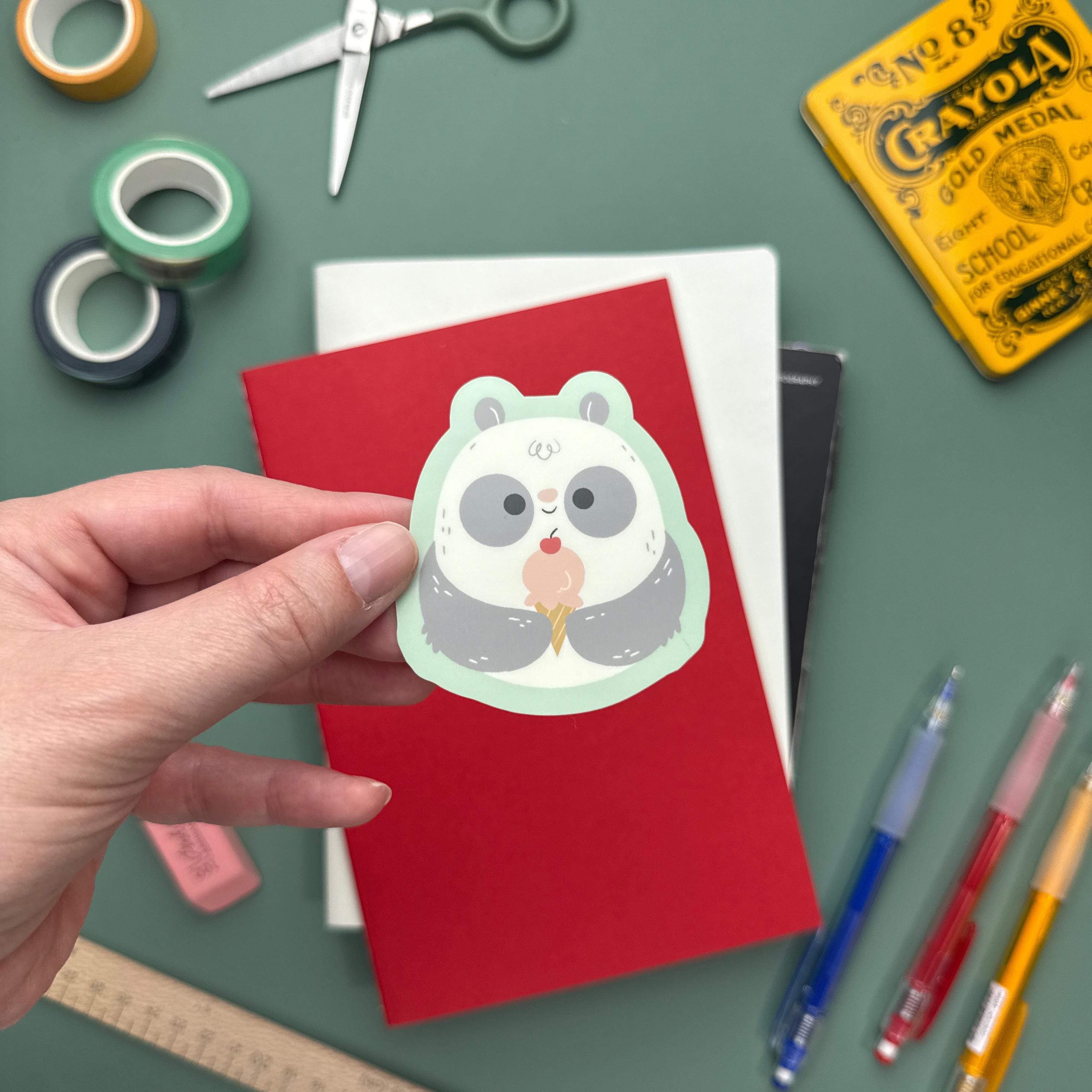 A hand holds a cute panda sticker featuring the panda eating an ice cream cone with a cherry on top. The background includes a red notebook, white journal, and various stationery items like washi tape, scissors, and pens.