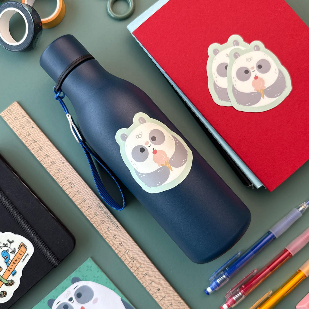 A navy blue water bottle decorated with a panda ice cream sticker sits on a green surface surrounded by school supplies, including notebooks, a wooden ruler, washi tape, and colorful pens.