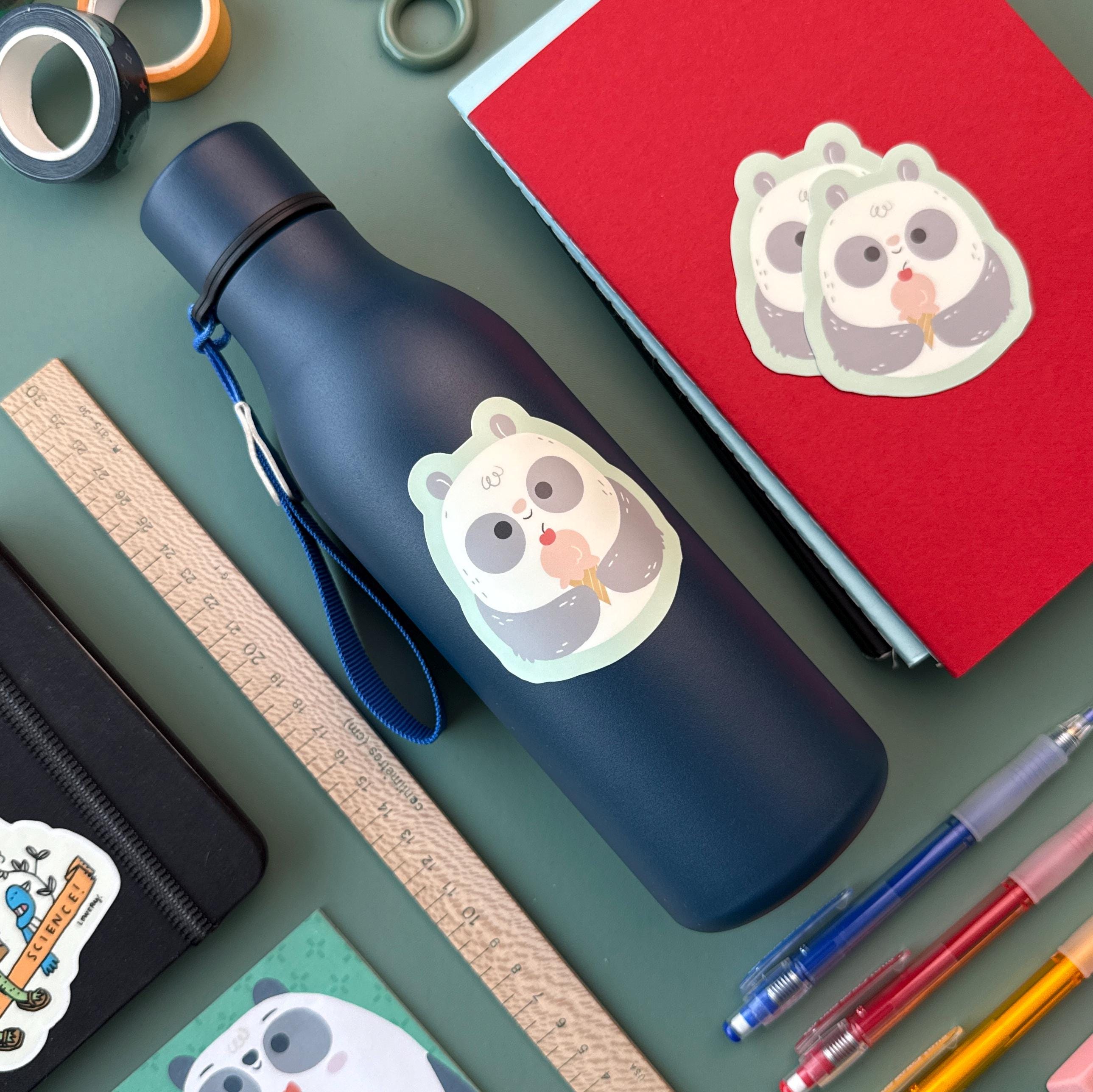 A navy blue water bottle decorated with a panda ice cream sticker sits on a green surface surrounded by school supplies, including notebooks, a wooden ruler, washi tape, and colorful pens.