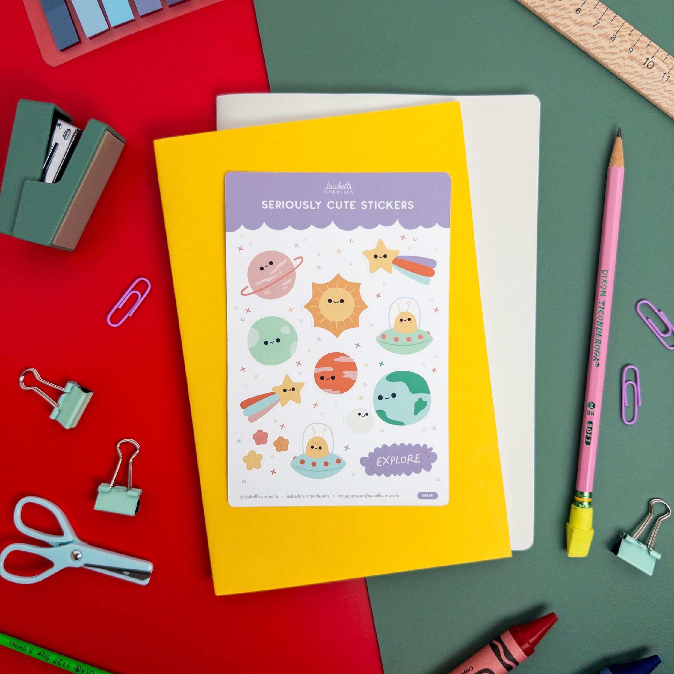 Kawaii planet-themed vinyl sticker sheet by Isabell’s Umbrella placed on a yellow notebook, surrounded by colorful stationery and art supplies.