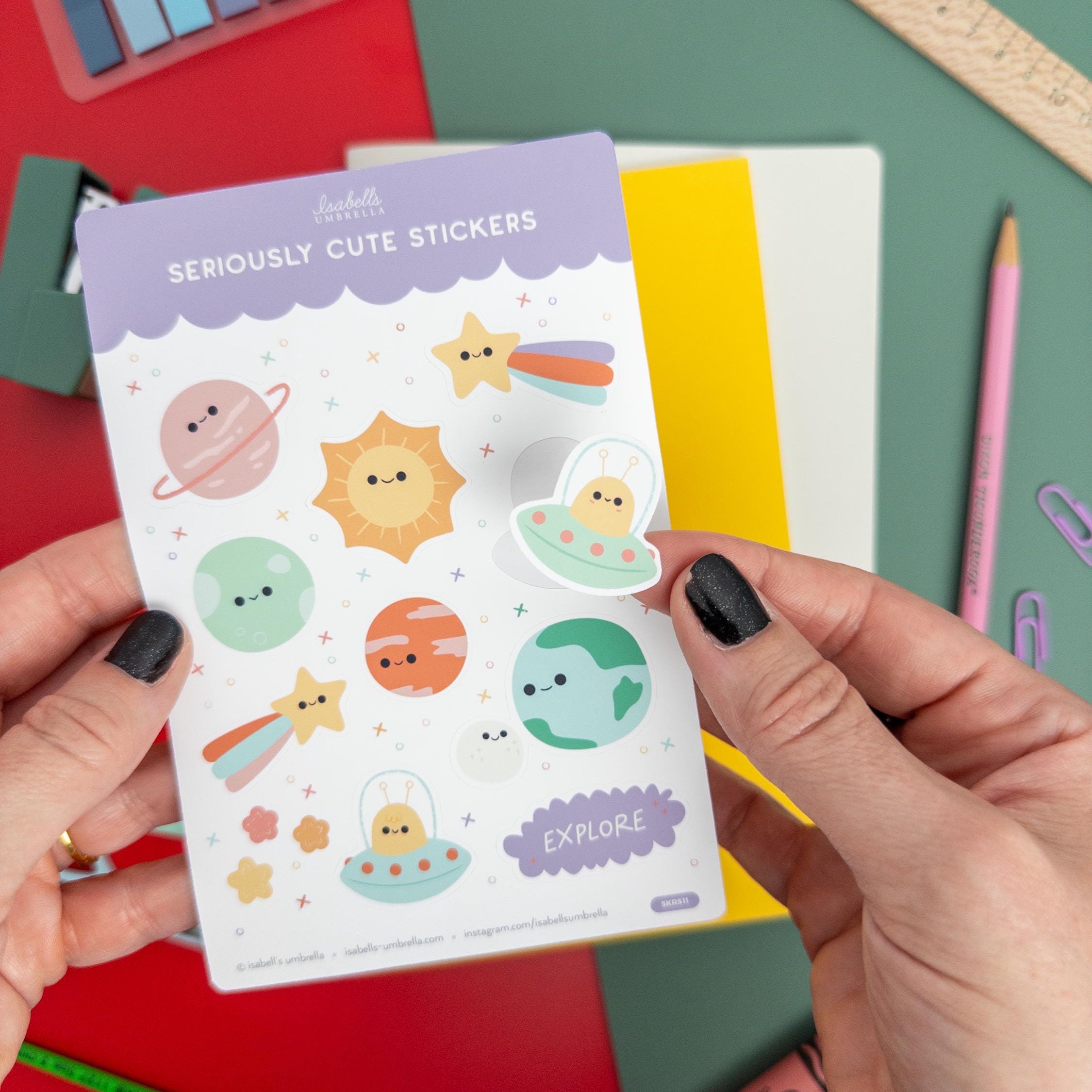 Hands holding a kawaii planet-themed vinyl sticker sheet featuring cute suns, planets, and stars from Isabell’s Umbrella on a colorful desk setup.