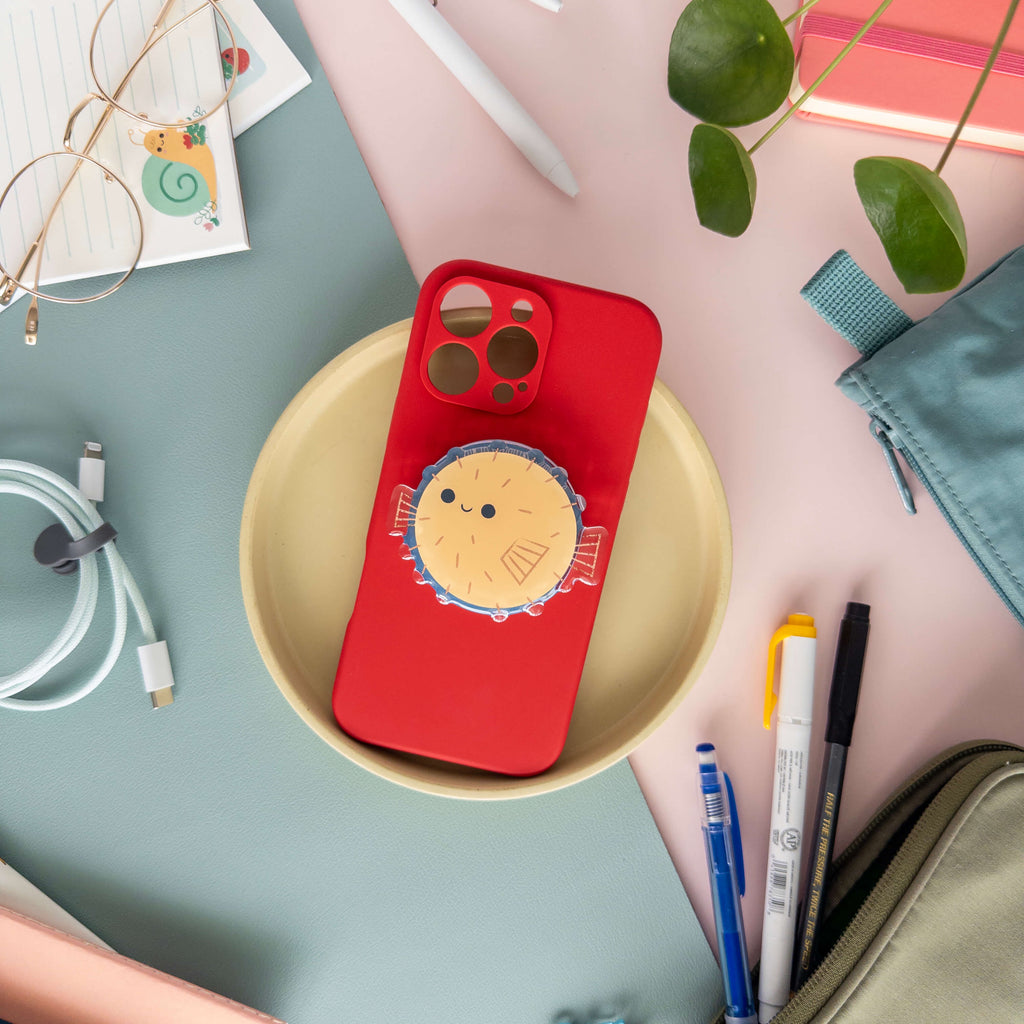 Red phone case with a magnetic phone grip of a pufferfish on a table with stationery. 