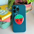 Blue phone case with a strawberry magnetic phone grip  on a white surface with books and plants.