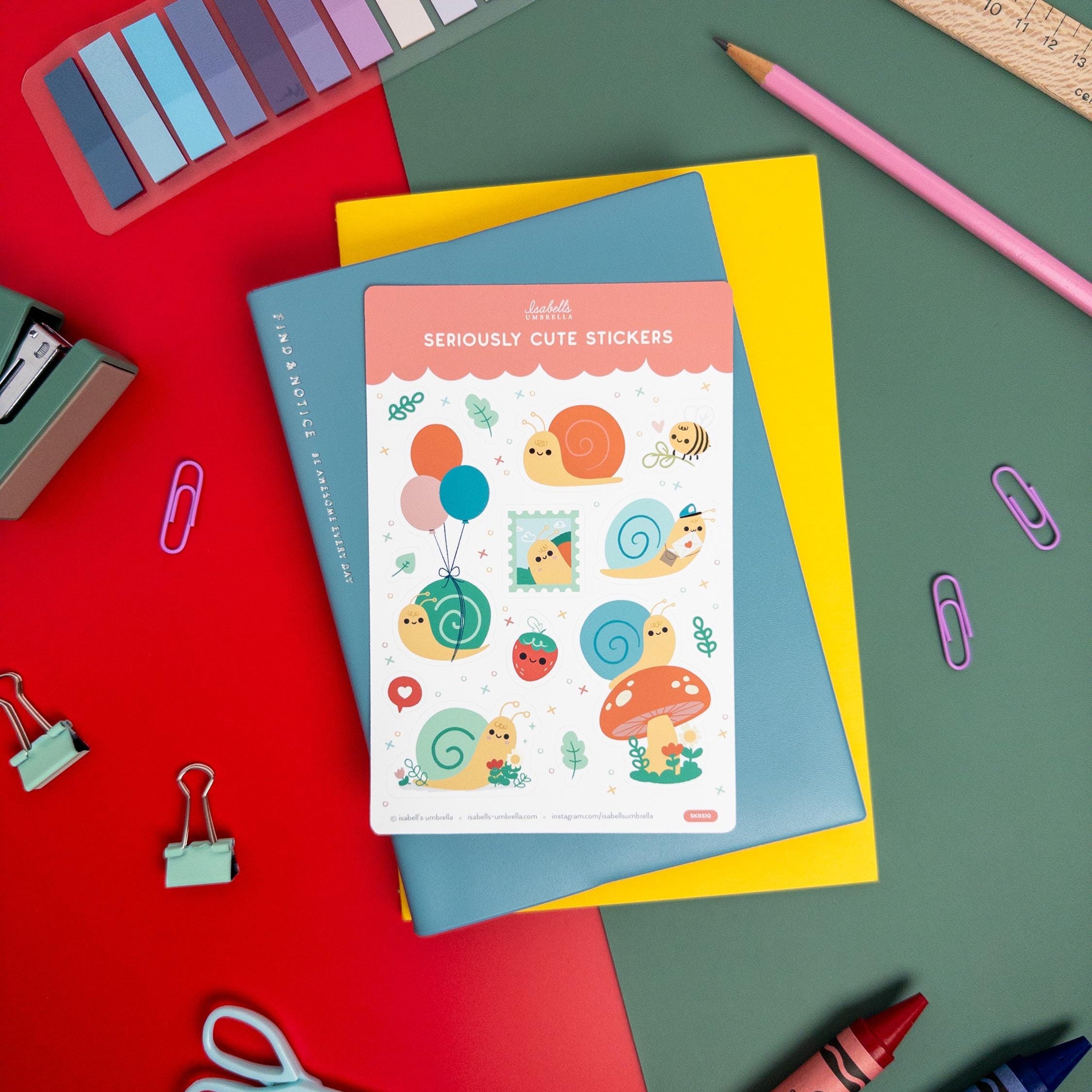 Kawaii snail-themed vinyl sticker sheet by Isabell’s Umbrella placed on a blue notebook, surrounded by colorful stationery and art supplies.