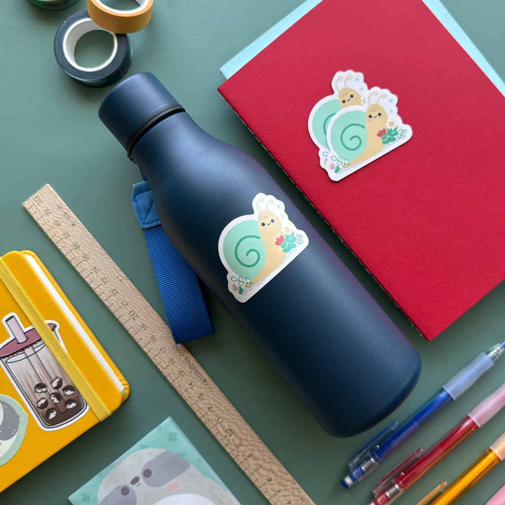 Cute snail surrounded by flowers sticker on a navy blue water bottle, surrounded by colorful pens, washi tape, a ruler, and notebooks. This waterproof vinyl sticker is perfect for decorating books, water bottles, laptops, and school supplies.