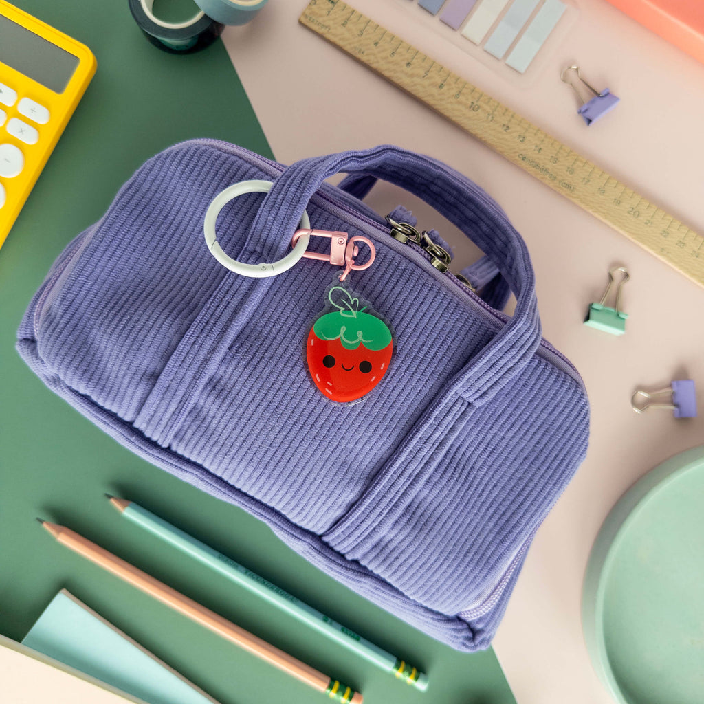 Purple pencil case with a strawberry keychain on a desk with stationery items