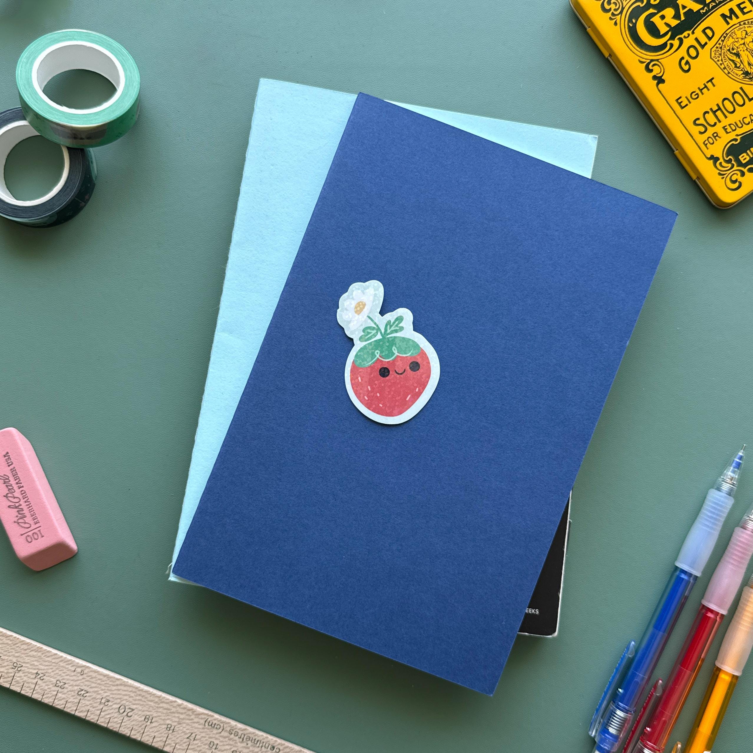 Kawaii strawberry with flower sticker applied to the cover of a blue notebook. The scene includes pens, a ruler, an erasure, and other stationery, highlighting this cute sticker as a fun addition to planners or journals.