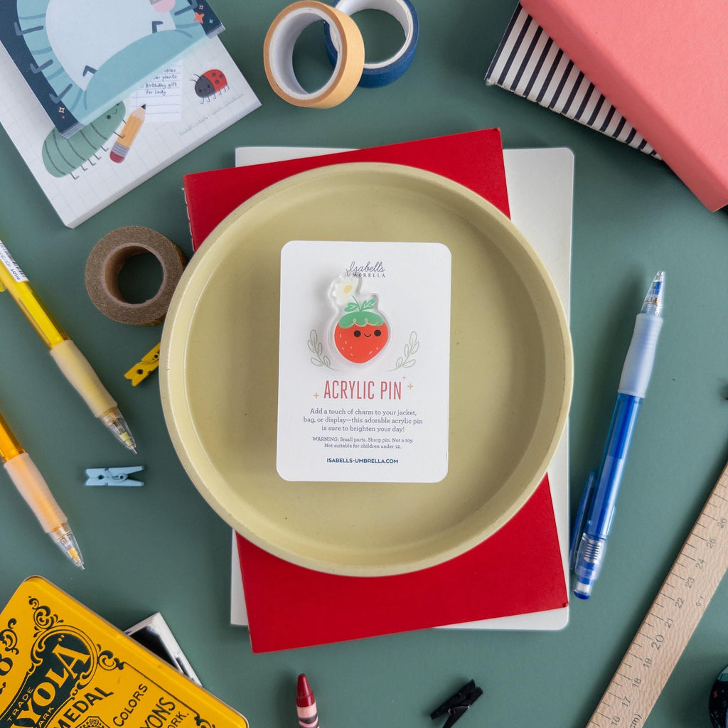A cute kawaii strawberry acrylic pin with flower detail sitting on yellow plate surrounded by colorful art supplies.
