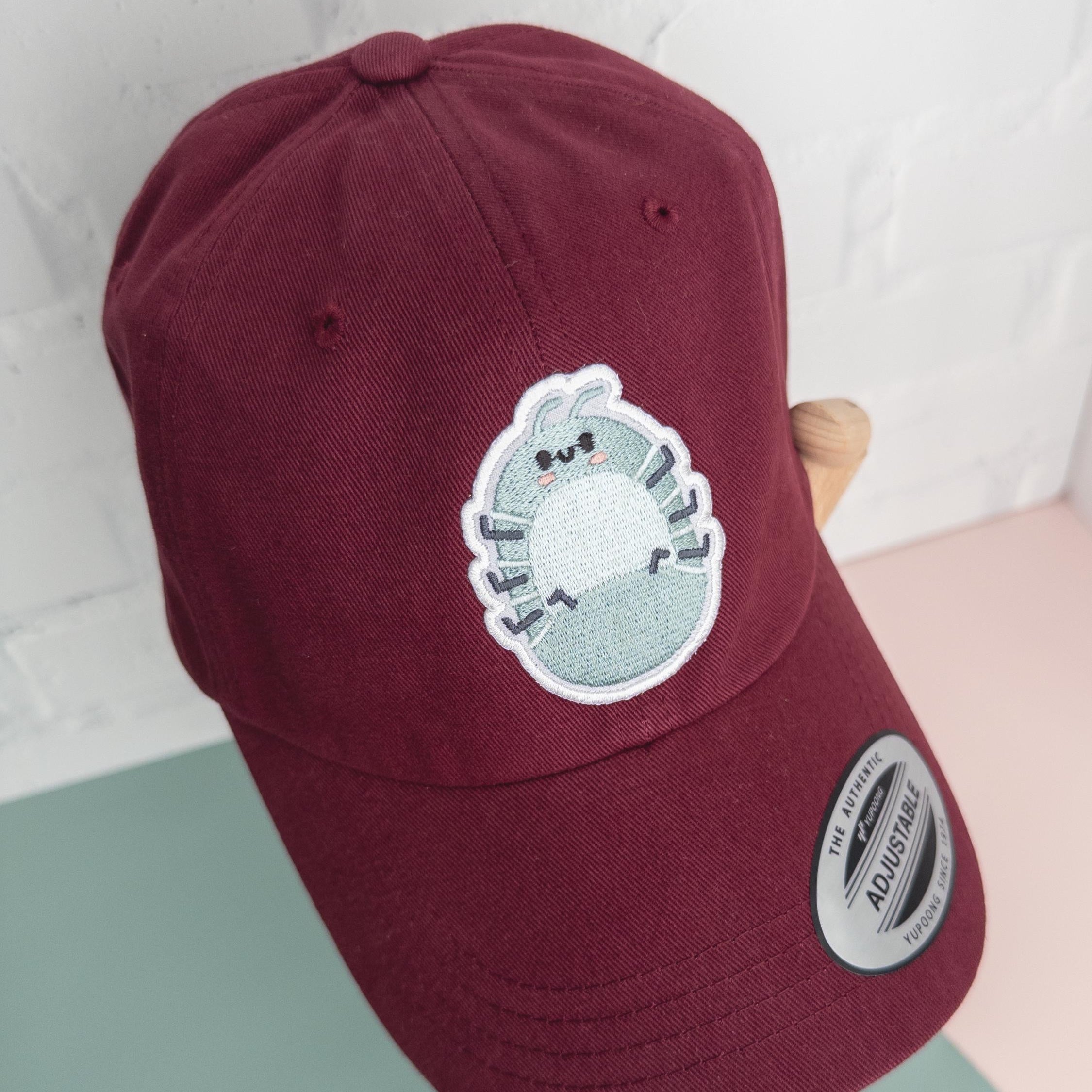 A maroon cotton twill dad hat with embroidered roly poly patch being held by artist wood hand model.