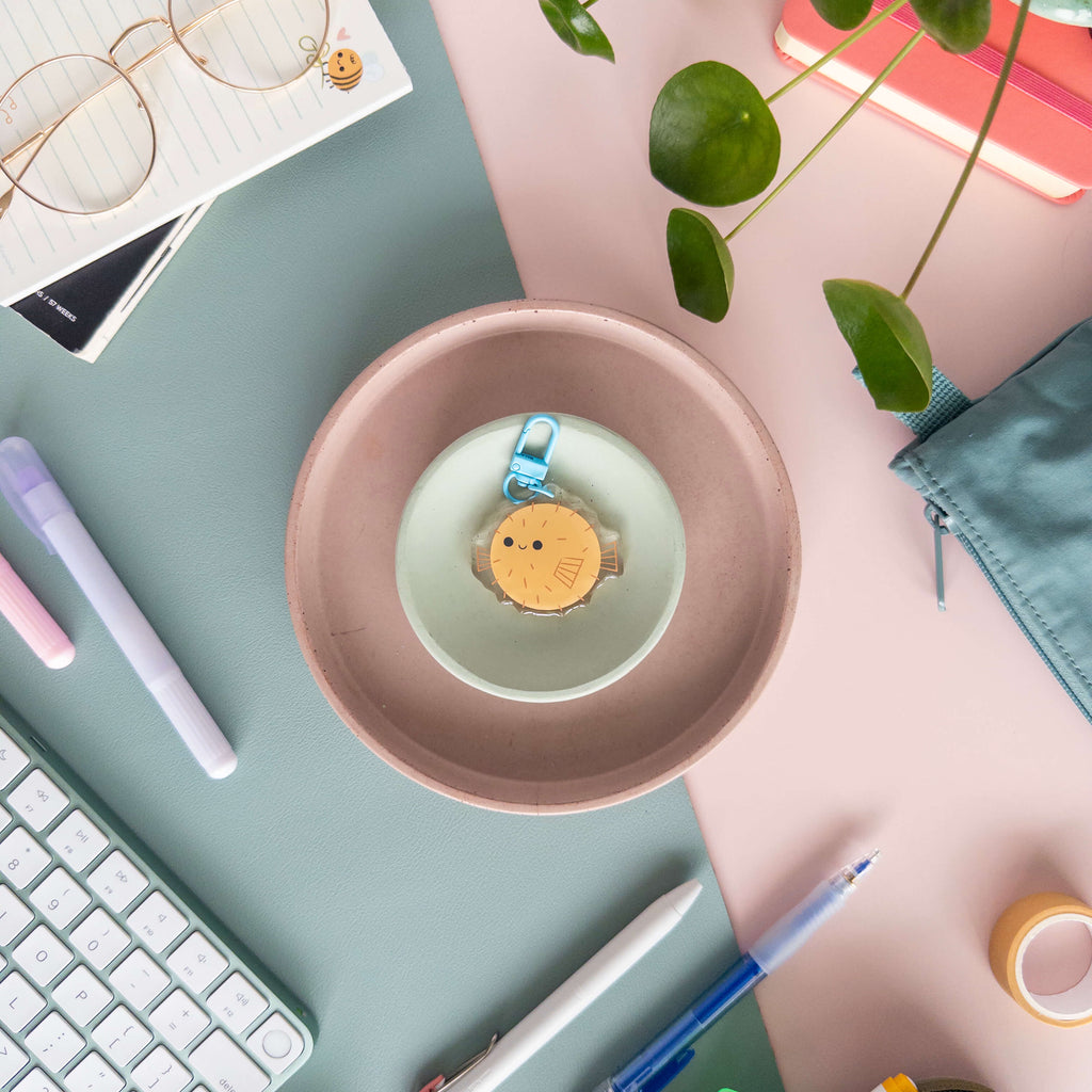 Desk setup with a pink bowl containing a small acrylic keychain of a yellow pufferfish .