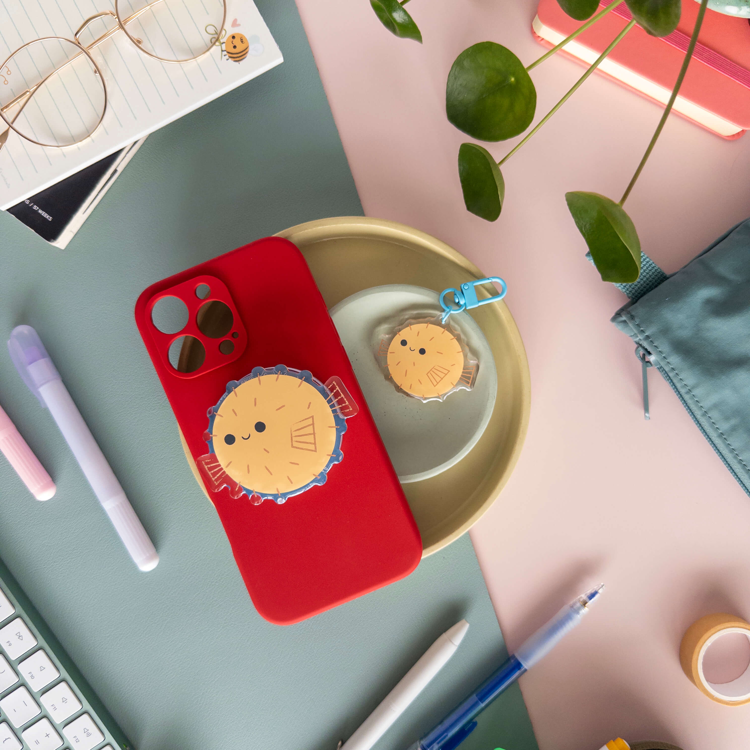 Red phone case with a yellow pufferfish magnetic phone grip next to a acrylic puffer fish keychain on a desk with stationery items