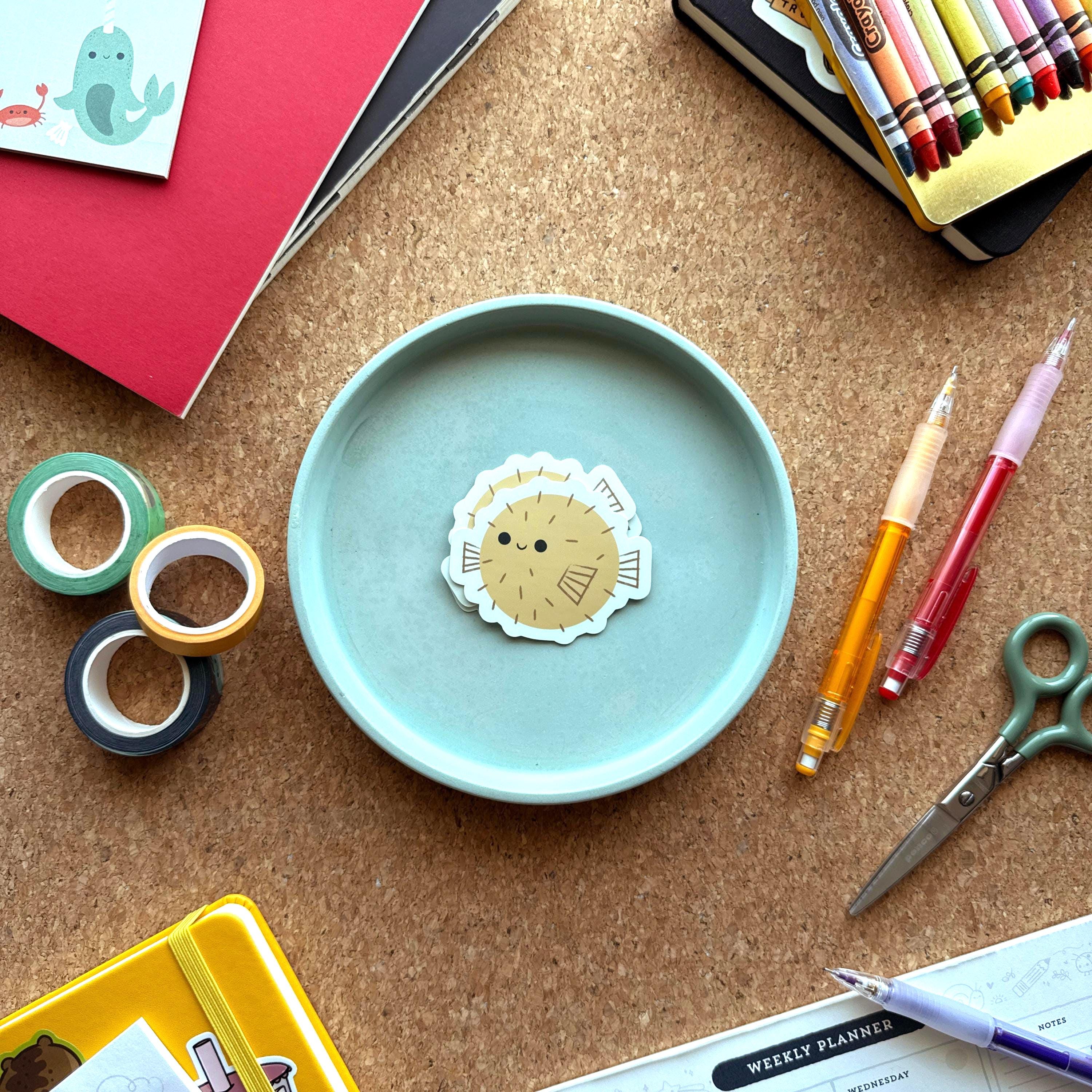 Kawaii sticker of a puffer fish set on top of a small blue tray. Surrounded by colorful pens, crayons, and notebooks.