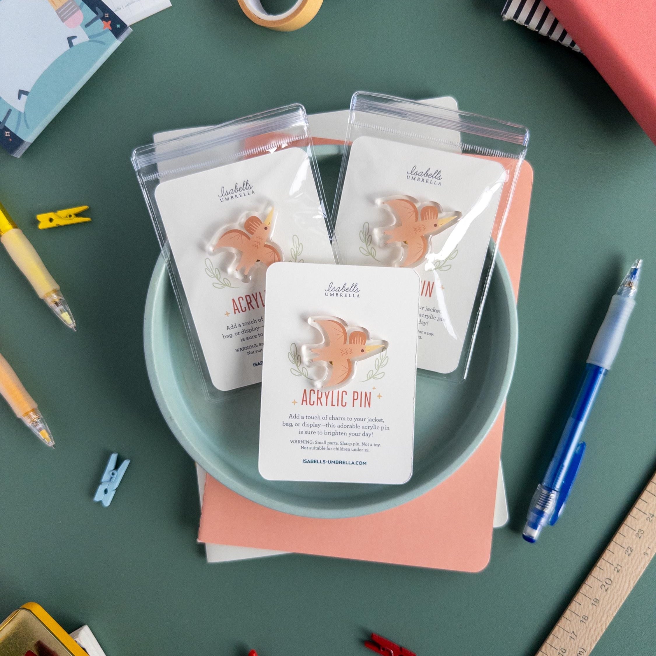 Three cute pterodactyl acrylic pin packaged and arranged on a blue plate with a variety of art supplies scattered around it.