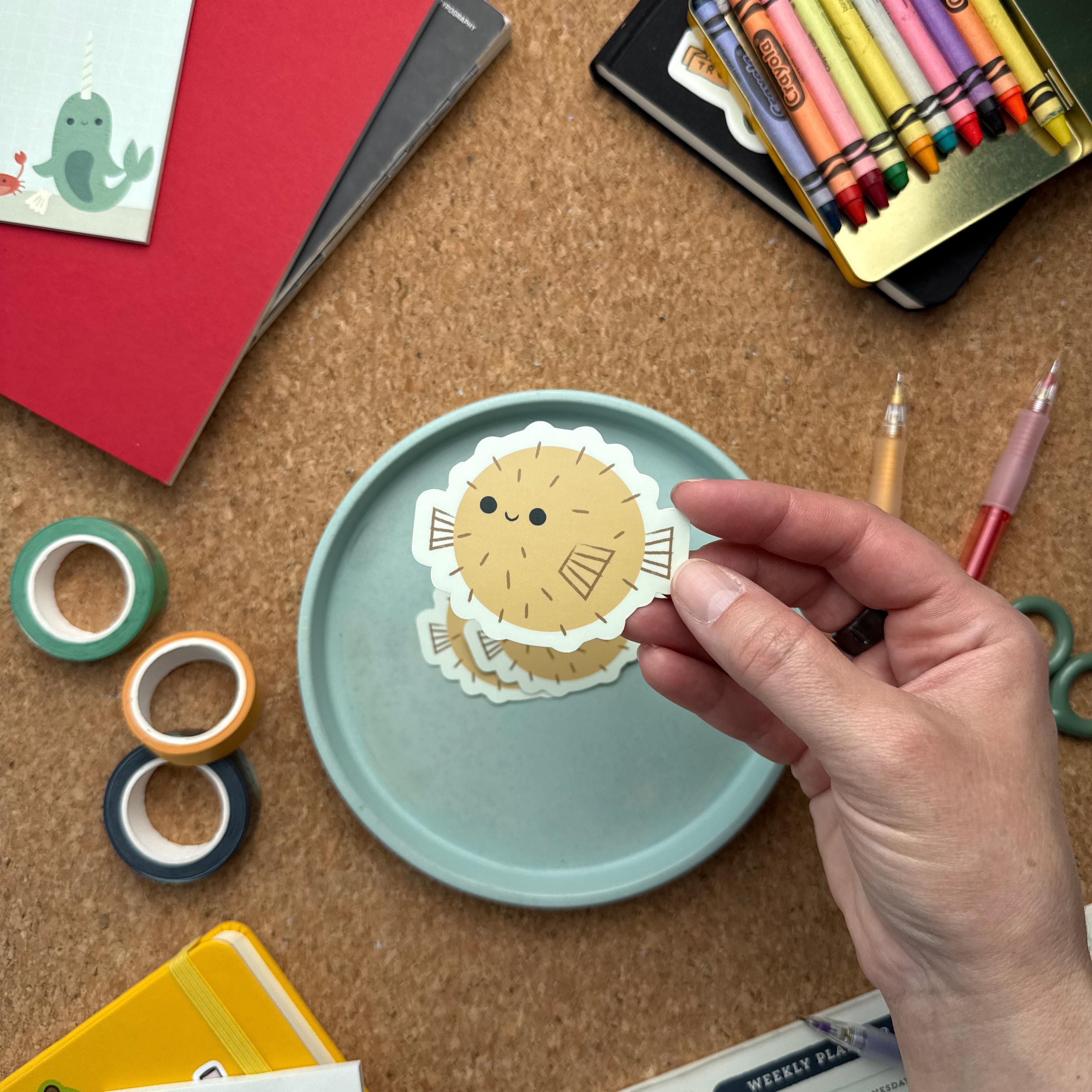 Hand holding an adorable puffer fish vinyl sticker. Surrounded by colorful stationery including washi tape, scissors, and notebooks—ideal for water bottles, journals, and planners.