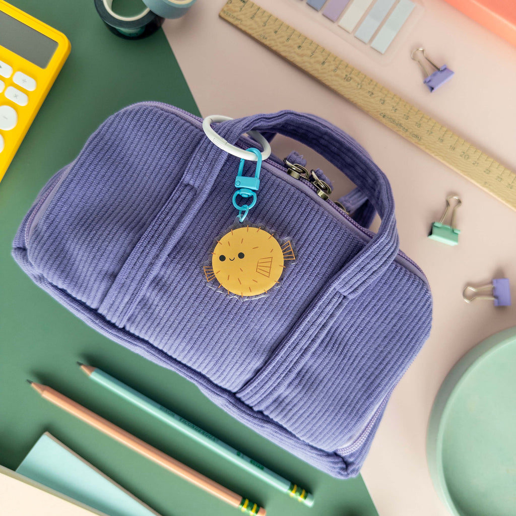 Purple corduroy pouch with a yellow pufferfish keychain on a desk with stationery items