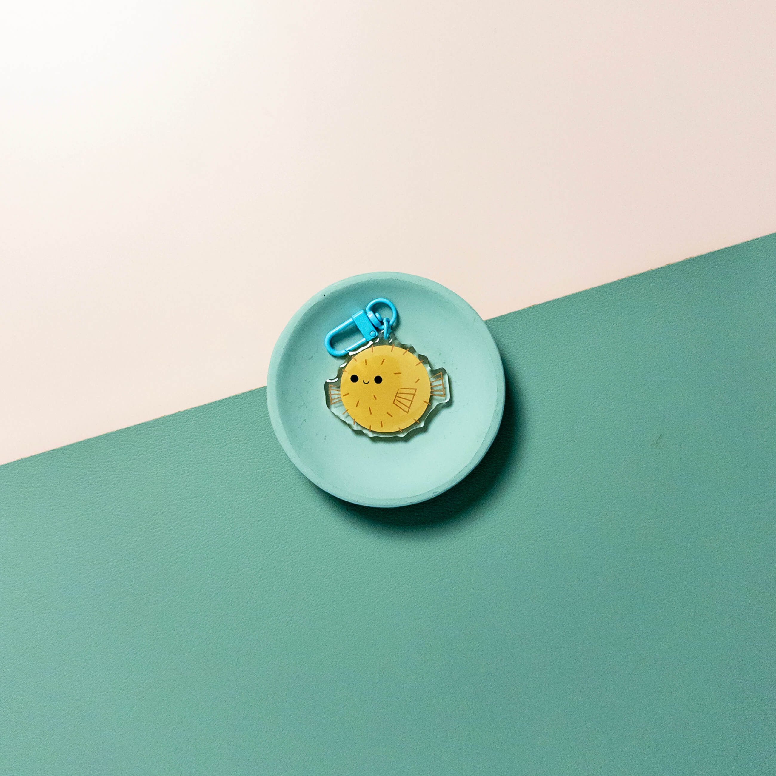 Small round teal dish with a yellow fish charm keychain on a green and beige background.
