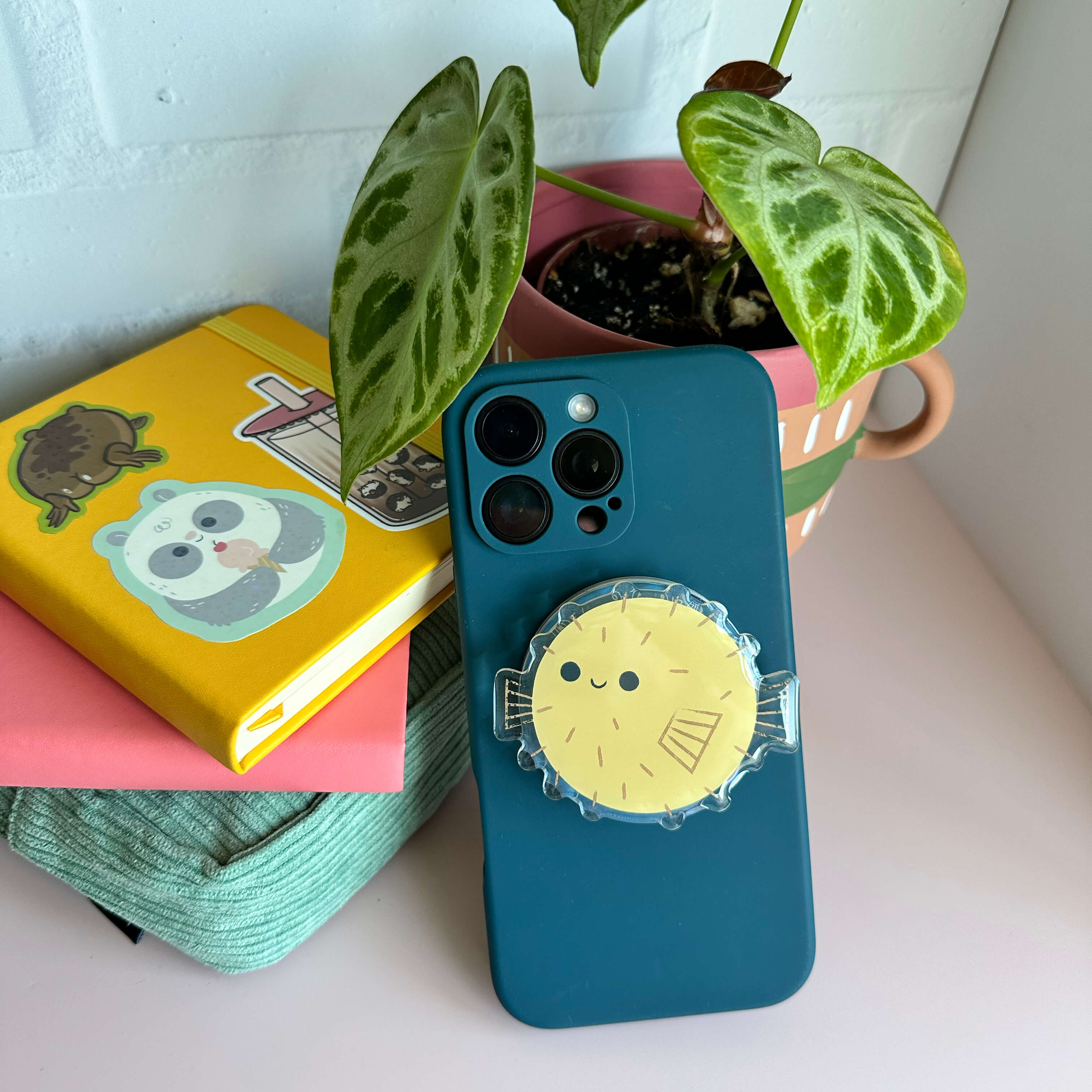 Phone with a blue case and yellow pufferfish magnetic phone grip on a surface with books and a plant in the background