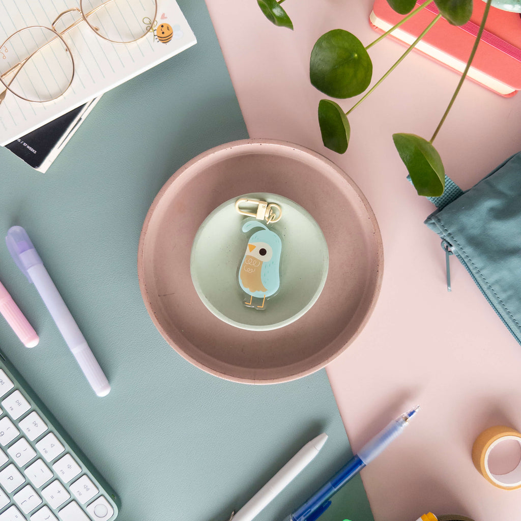 Desk setup with a pink bowl containing a small acrylic quail keychain , surrounded by stationery items.