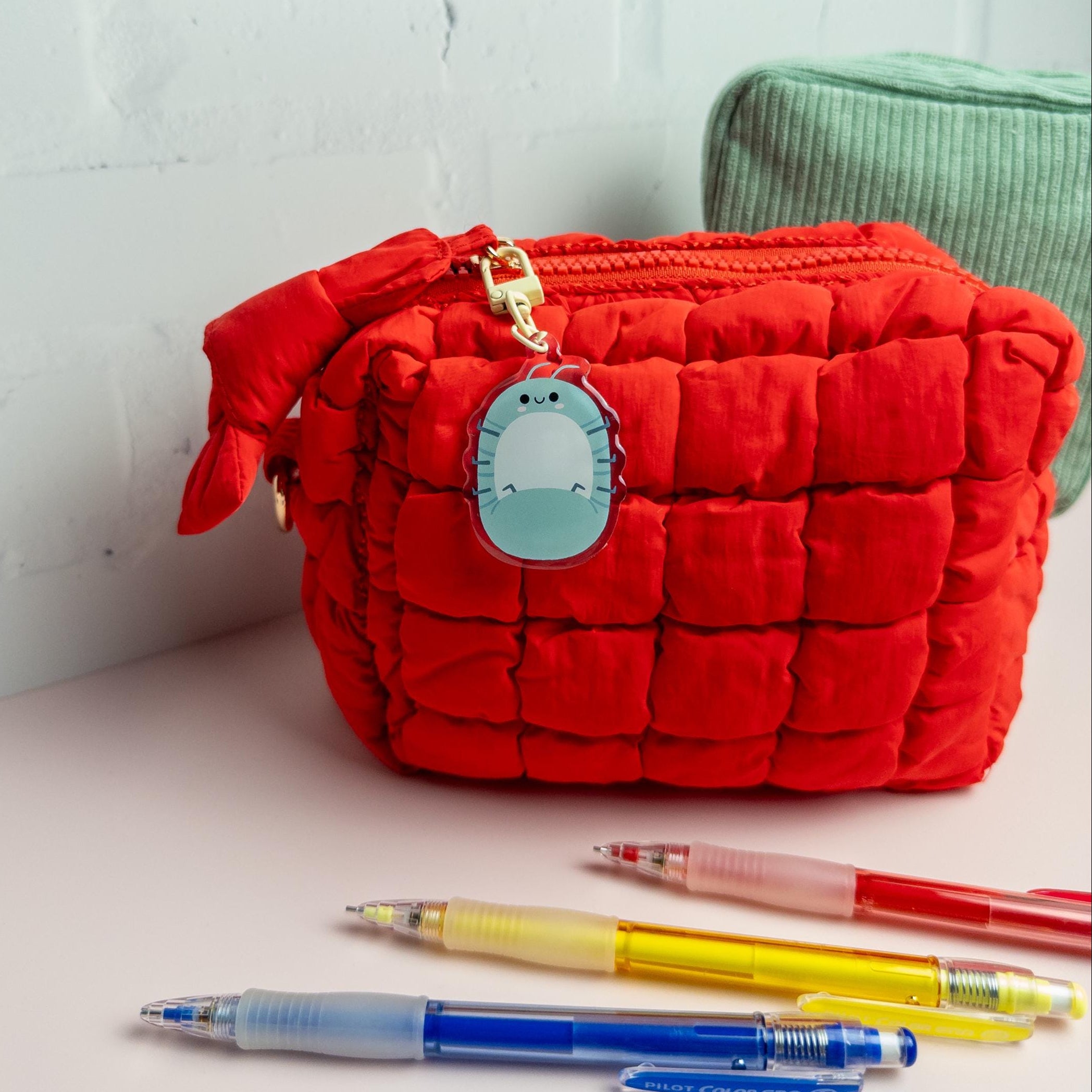 Red quilted pouch with a gold zipper and blue emblem on a light surface with pens.