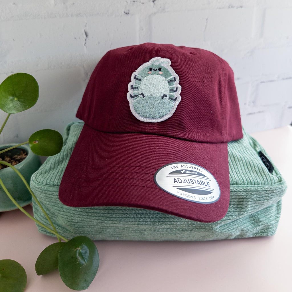 A maroon cotton twill dad hat with embroidered roly poly patch sitting on top of a sage green bag with plant sitting beside it.