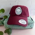 A maroon cotton twill dad hat with embroidered roly poly patch sitting on top of a sage green bag with plant sitting beside it.