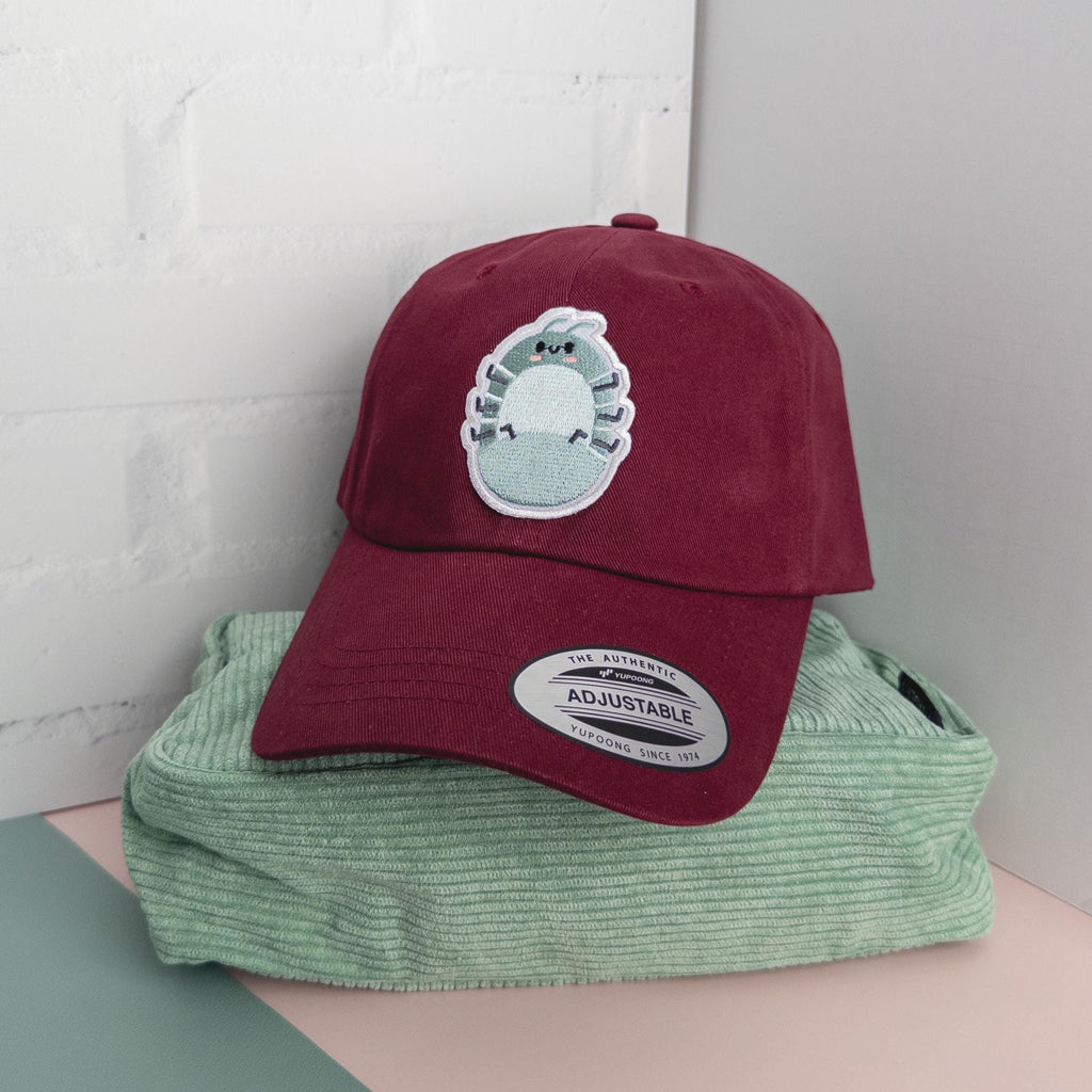 A maroon cotton twill dad hat with embroidered roly poly patch sitting on a mint green pouch.