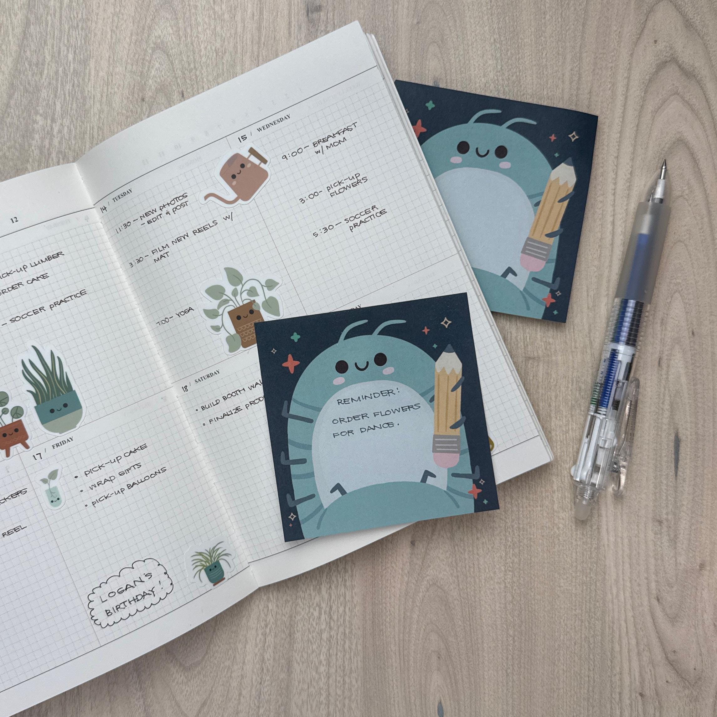 Sticky notepad featuring a roly poly holding a pencil. The notepad is sitting on a a calendar, with a pen sitting beside it.