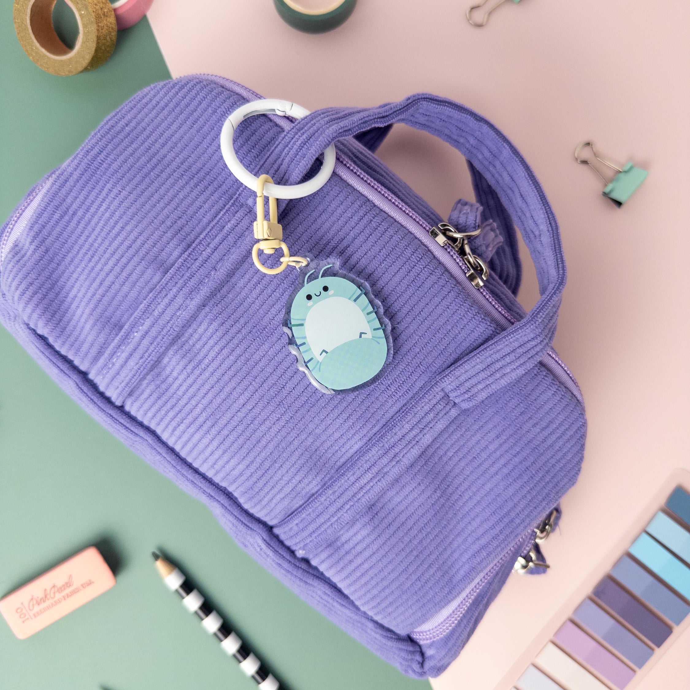 .Purple bag with a Roly Poly bug keychain clipped to it on a colorful background with stationery items