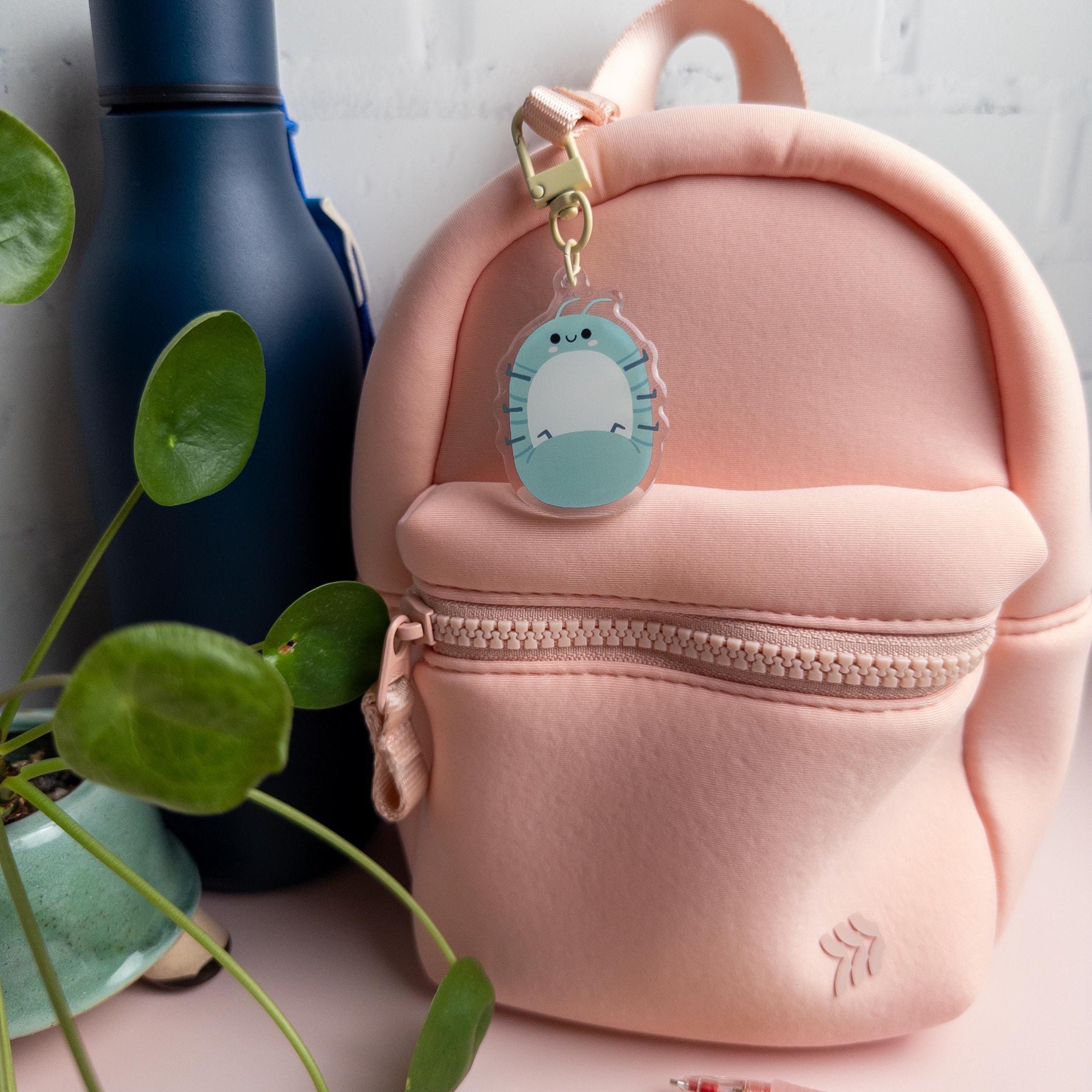 Pink backpack with a roll poly keychain clipped to the zipper and pictured on a surface with pens and a plant