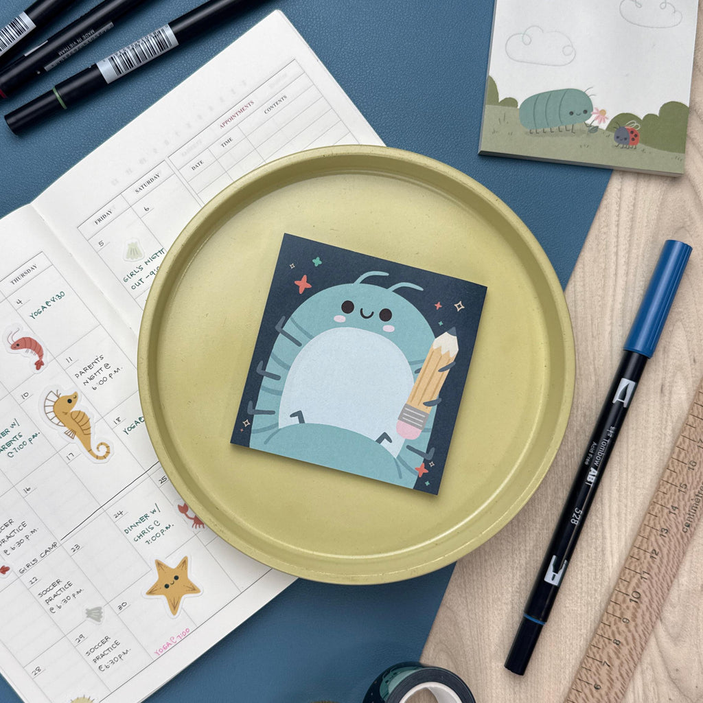 Sticky notepad featuring a roly poly holding a pencil. The notepad is sitting on a yellow plate and is surrounded by a calendar, markers, ruler, and another notepad.