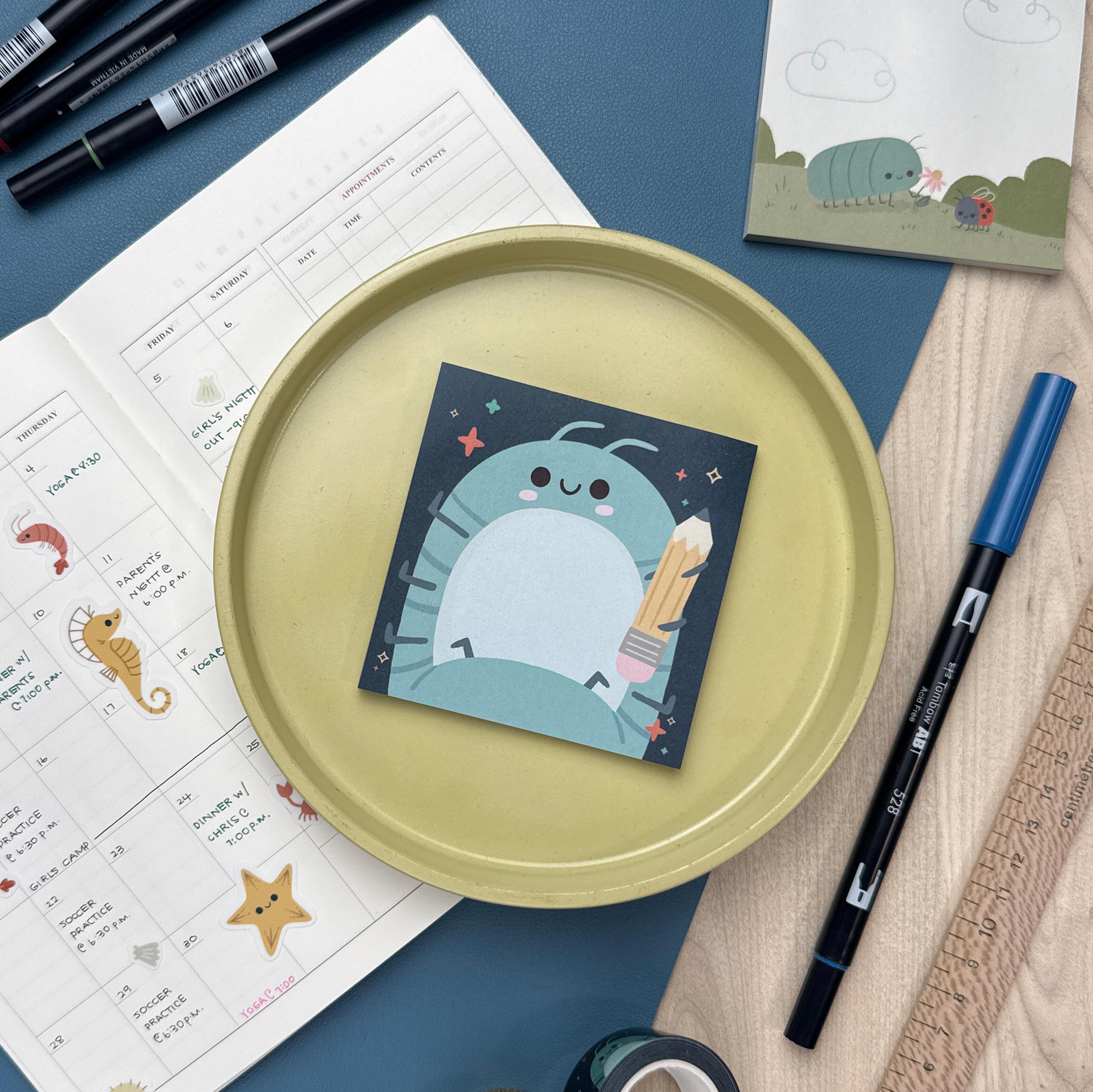Sticky notepad featuring a roly poly holding a pencil. The notepad is sitting on a yellow plate and is surrounded by a calendar, markers, ruler, and another notepad.
